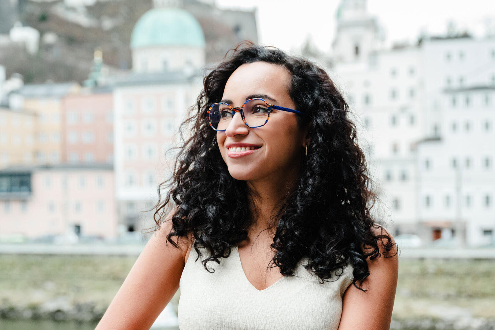 Eine Frau mit lockigem dunklem Haar und Brille lächelt, während sie im Freien in einer Stadt mit historischen Gebäuden und einem Kuppelbau im Hintergrund steht.