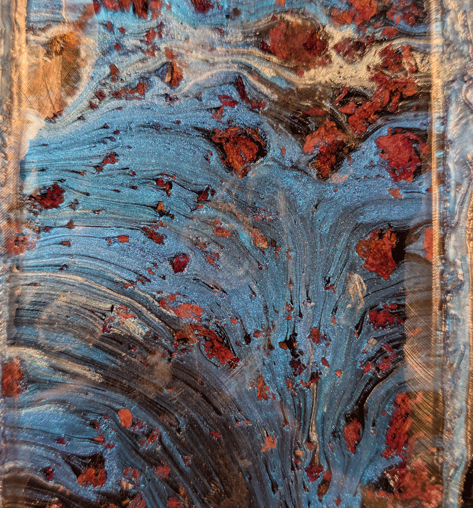 Wirbel aus schimmernder blauer Farbe mit verstreuten rötlich-braunen Materialbrocken ergeben ein strukturiertes, abstraktes Muster, das an fließendes Wasser und verstreute Steine erinnert.