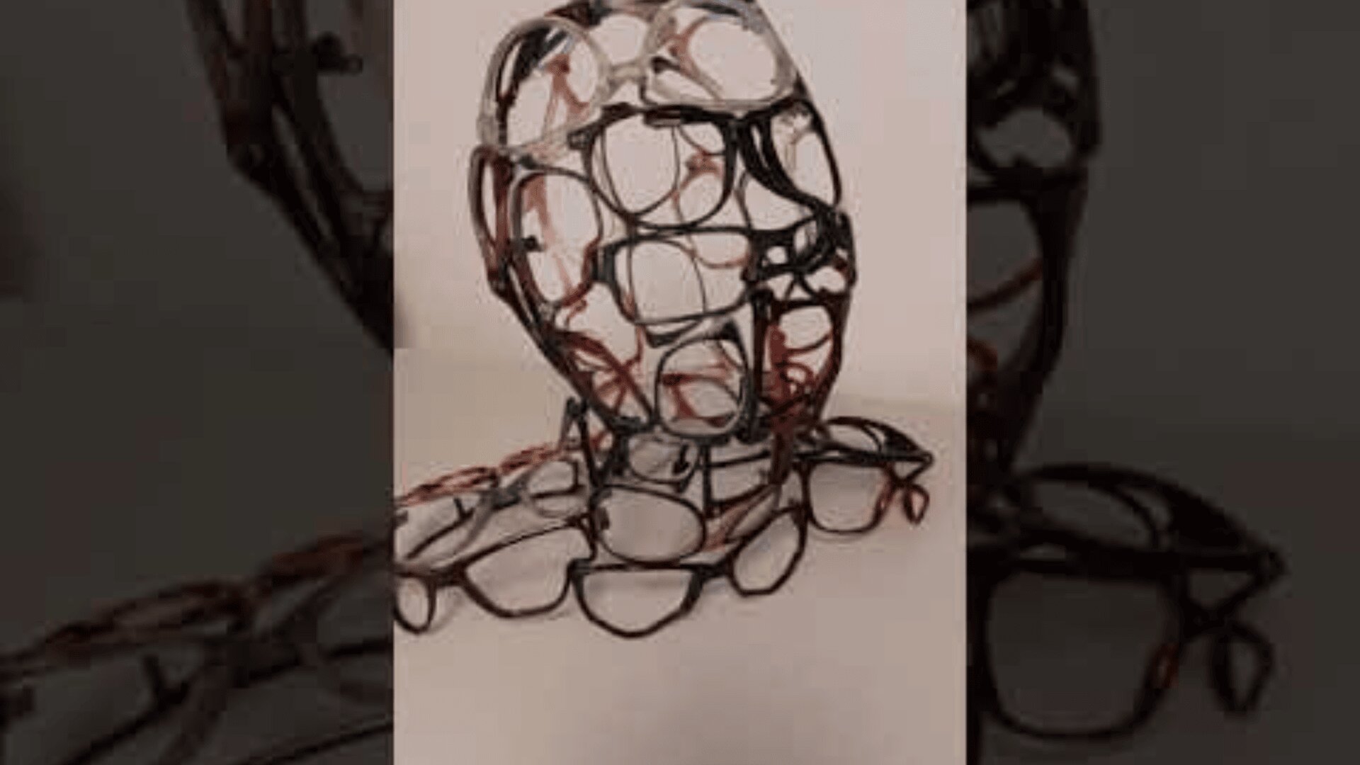 Eine Skulptur eines menschlichen Kopfes und von Schultern, die vollständig aus miteinander verbundenen Brillengestellen besteht und vor einem schlichten Hintergrund präsentiert wird.