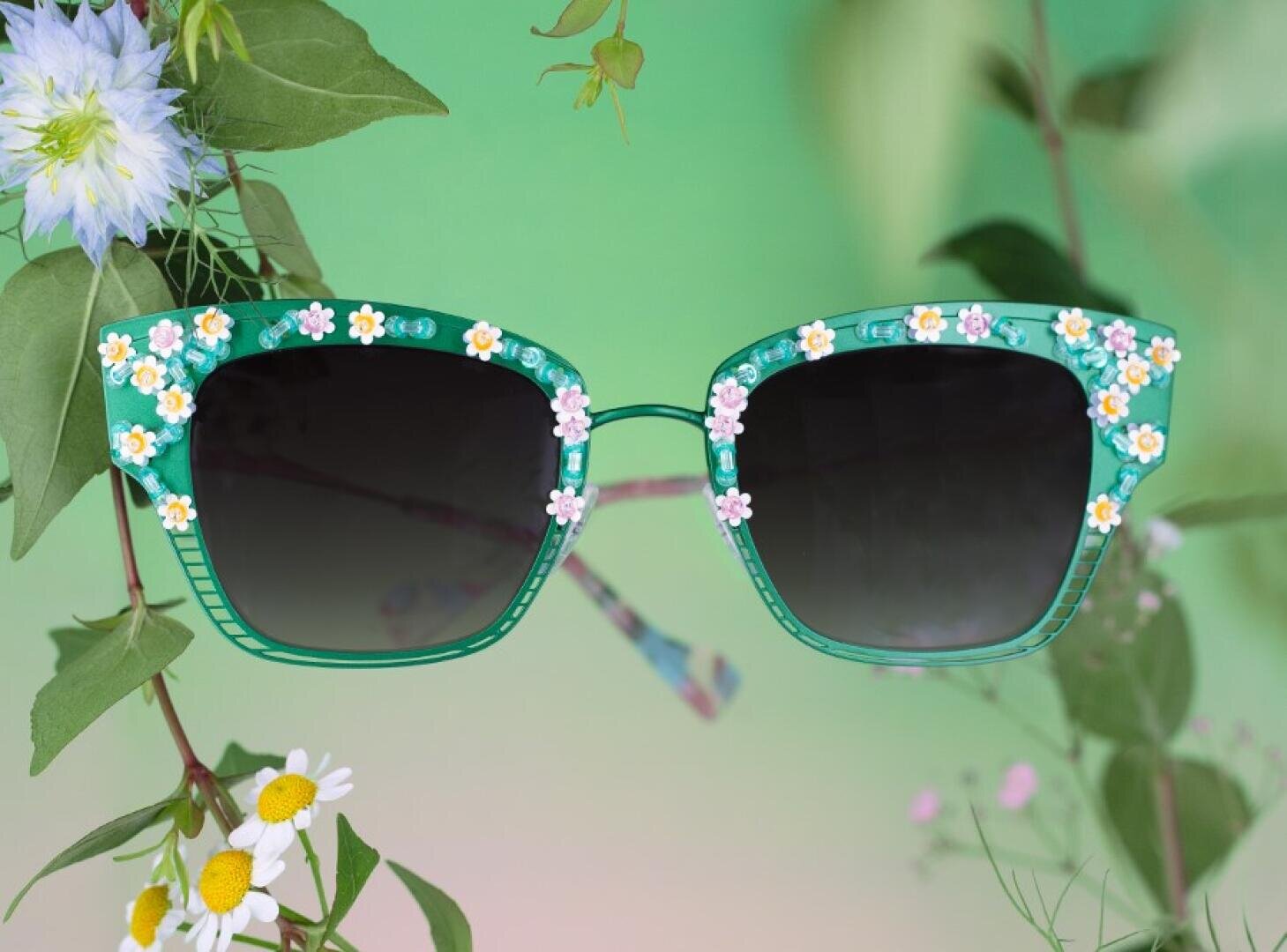 Eine grüne Sonnenbrille mit kleinen weißen und gelben Blüten hängt zwischen belaubten Zweigen und blühenden Blumen vor einem zartgrünen Hintergrund.