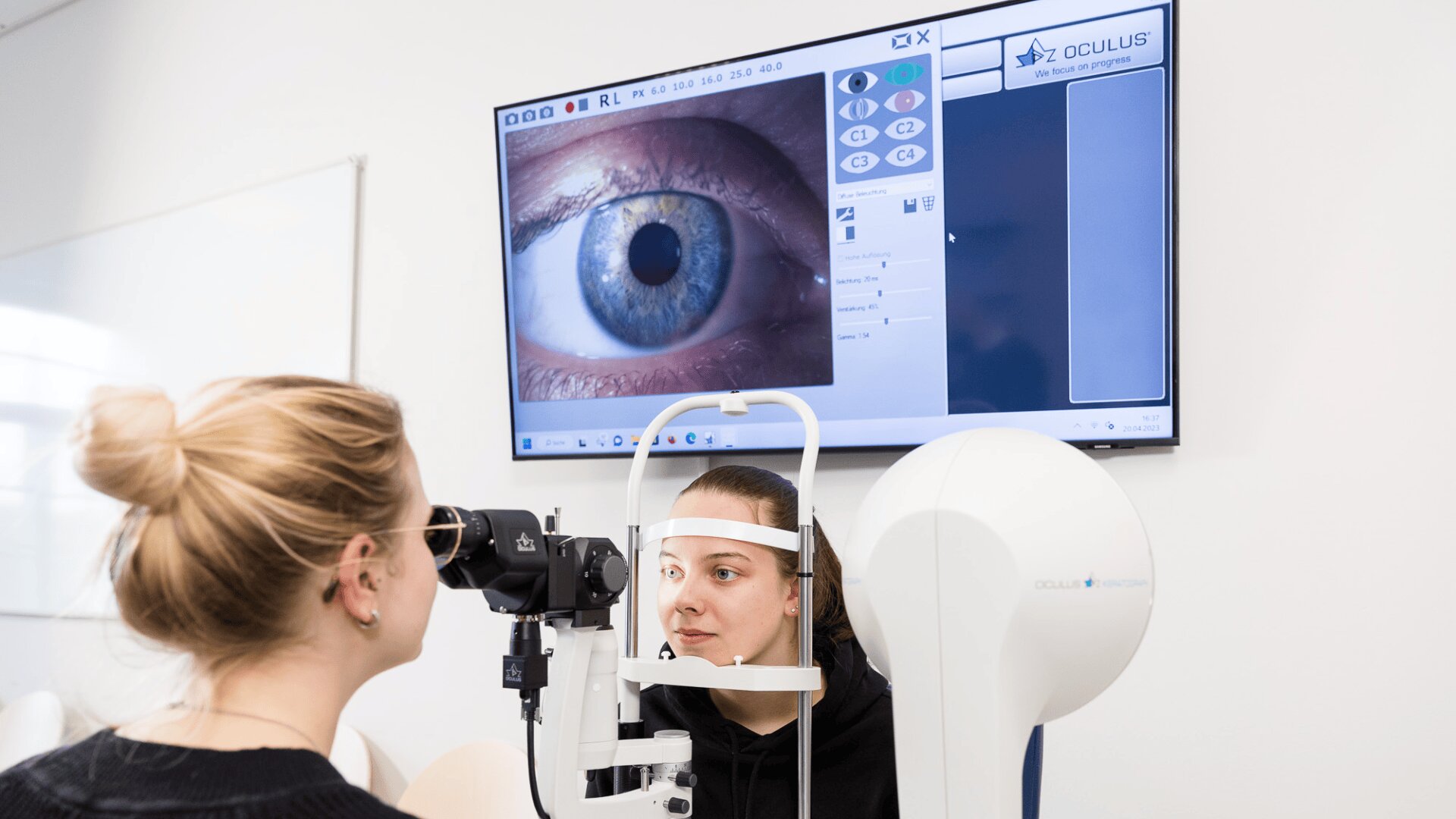 Eine Frau sitzt an einem Augenuntersuchungsgerät, während ein Optometrist einen Sehtest durchführt. Ein großer Monitor zeigt eine Nahaufnahme des Auges des Patienten und diagnostische Informationen in einer klinischen Umgebung an.