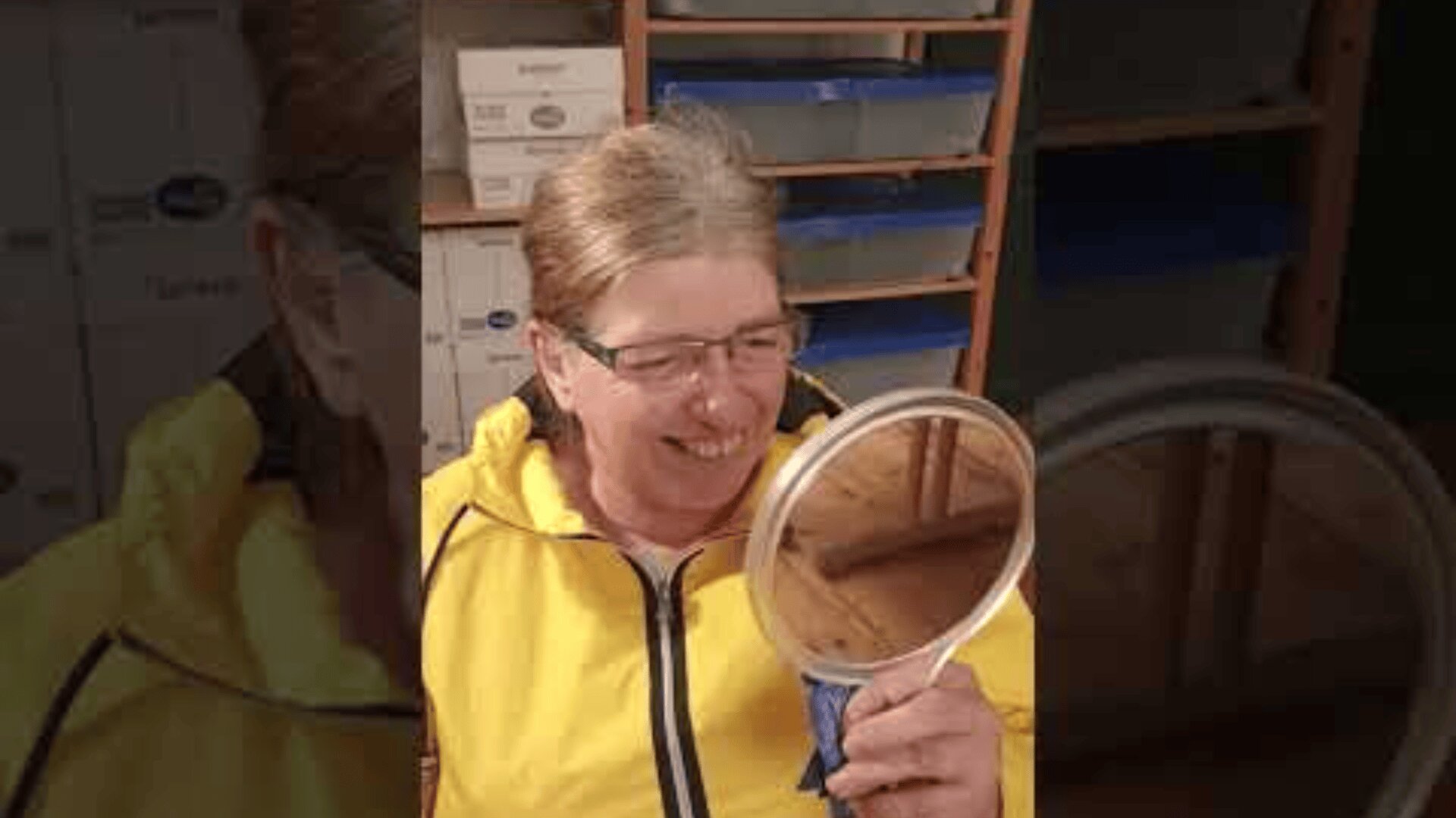 A smiling person with glasses and a yellow jacket looks at themselves in a handheld round mirror, sitting indoors with shelves and boxes in the background.