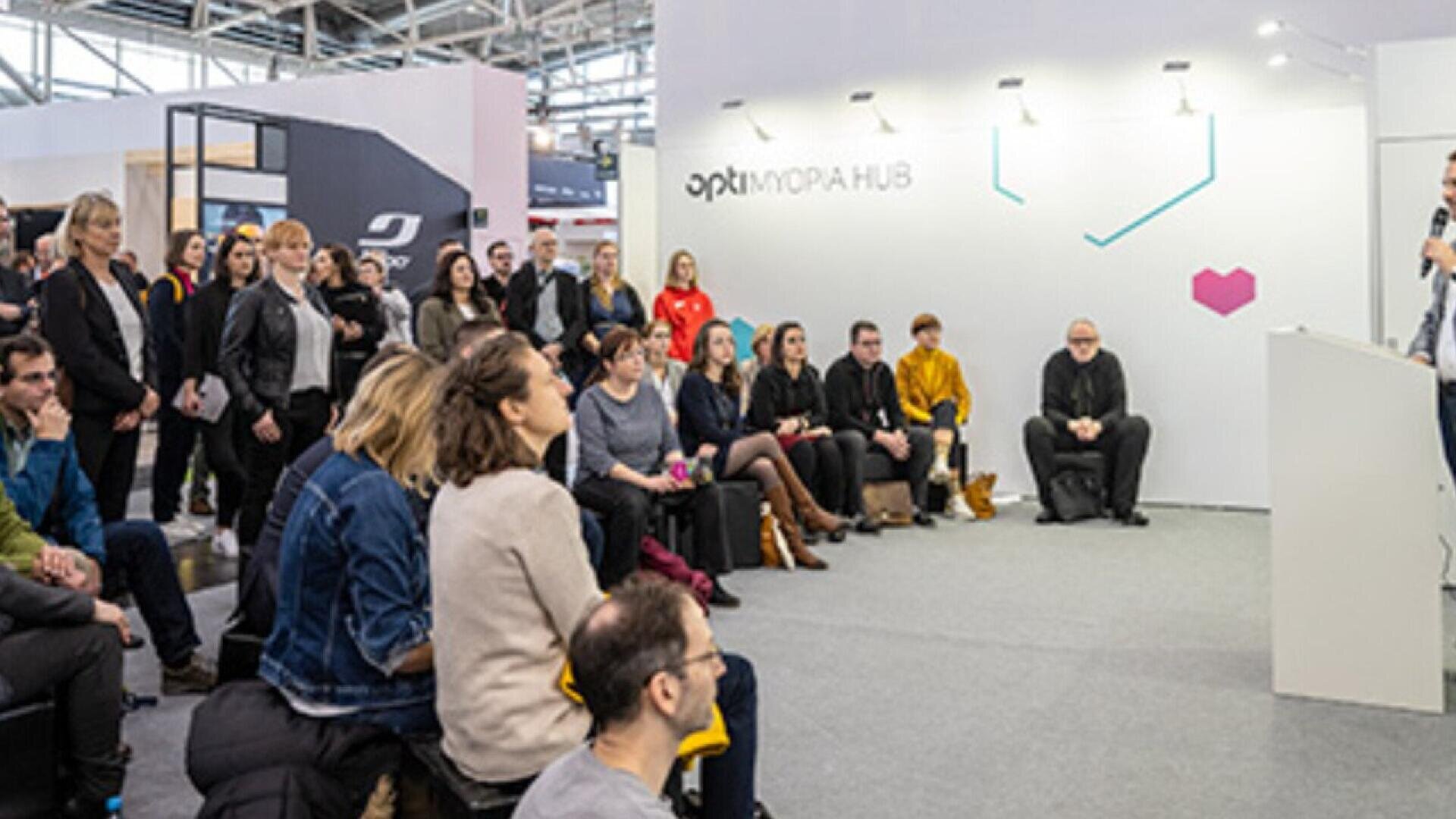 A group of people sit and stand attentively, listening to a speaker at a modern indoor event space labeled “OPTI MORPH HUB.” The setting appears professional and focused.