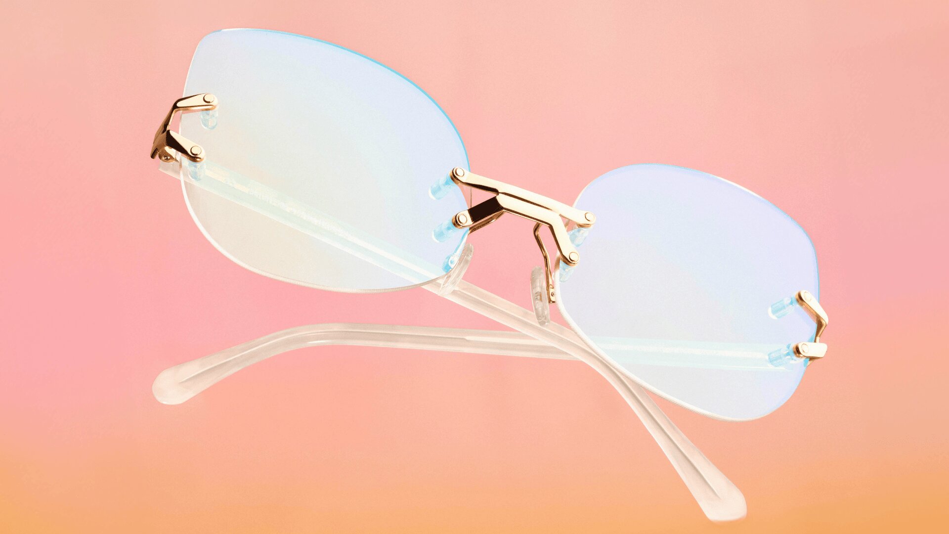 Eine randlose Brille mit goldenen Scharnieren und klaren, leicht getönten blauen Gläsern, ausgestellt auf einem transparenten Gestell vor einem Hintergrund mit rosa und orangefarbenem Farbverlauf.