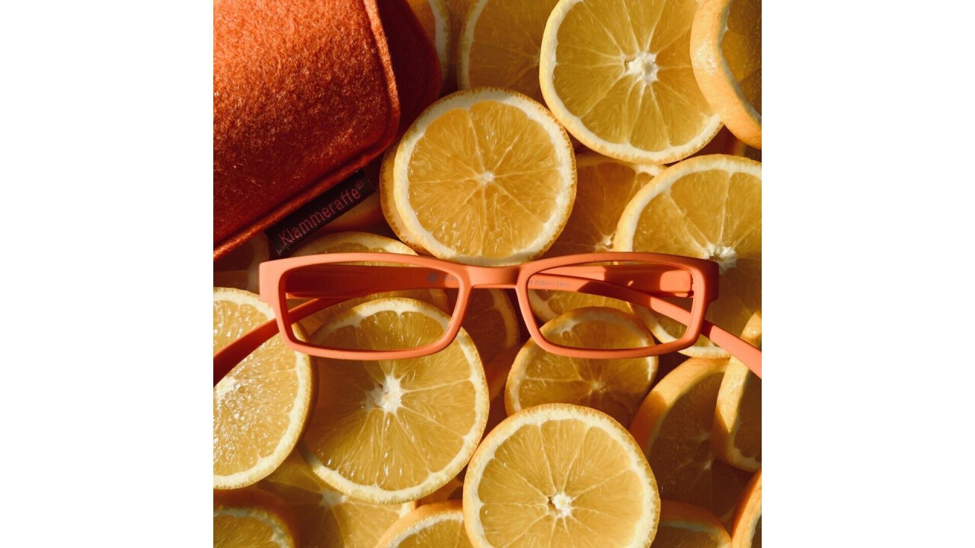 A pair of rectangular orange glasses and an orange glasses case rest on top of a pile of orange slices, blending in with the citrus background.
