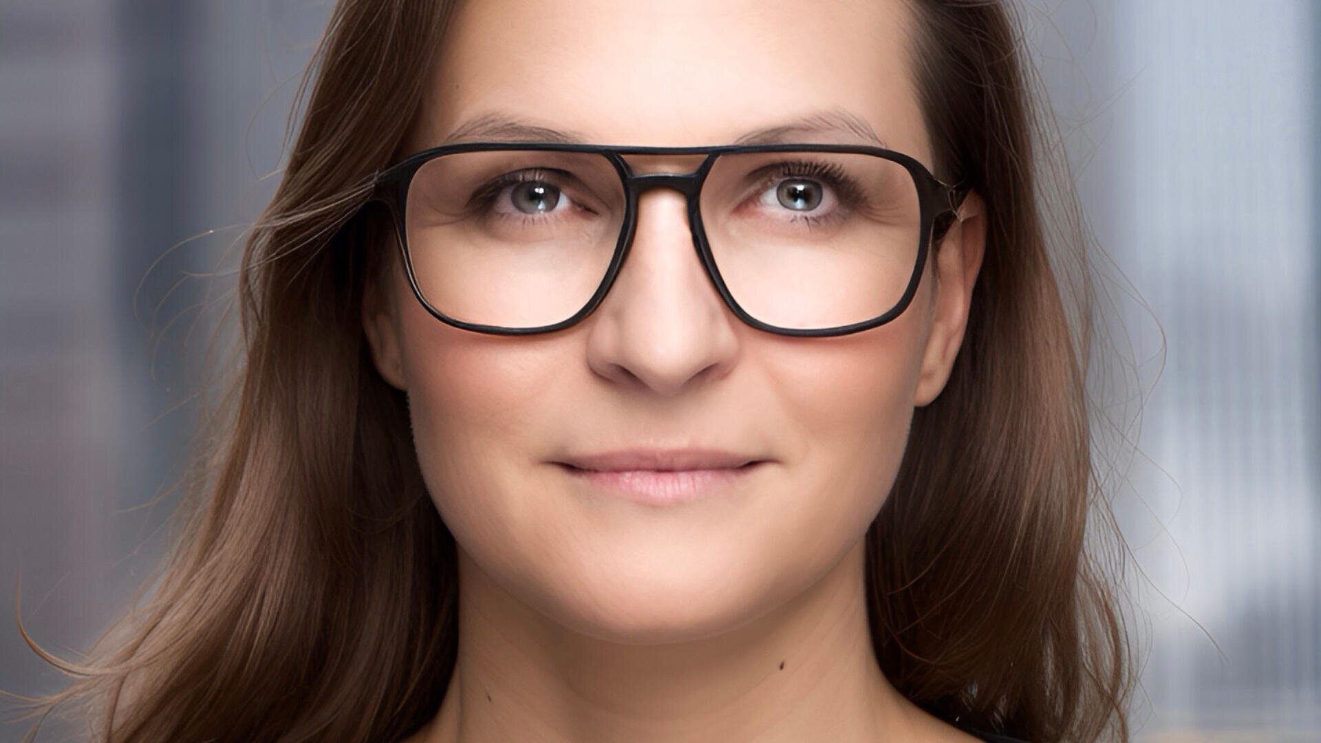 Eine Frau mit langen braunen Haaren und einer großen schwarzen Brille lächelt sanft. Der Hintergrund ist unscharf und lässt auf eine Innen- oder Büroumgebung schließen.
