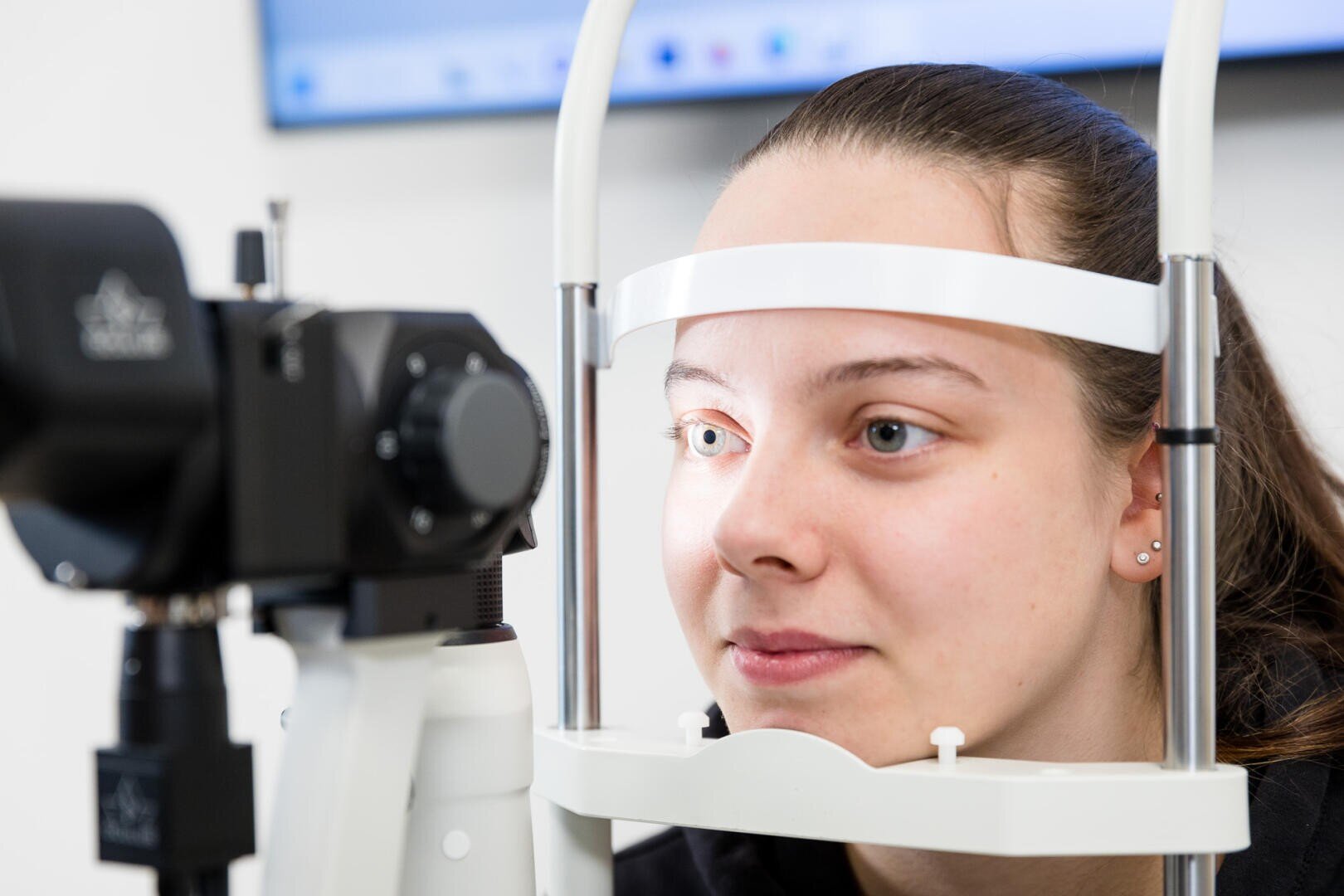 Eine junge Frau unterzieht sich einer Augenuntersuchung, wobei sie ihr Kinn und ihre Stirn auf eine Stütze stützt, während sie in ein ophthalmisches Gerät schaut, wahrscheinlich in einer Augenarztpraxis.