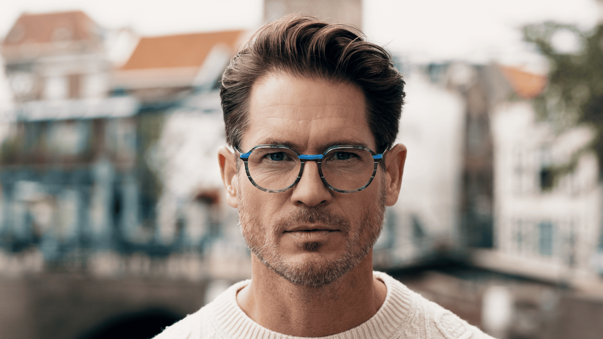 Ein Mann mit kurzen braunen Haaren und einer blauen Brille, der einen weißen Pullover trägt, steht im Freien mit unscharfen Gebäuden und Grünflächen im Hintergrund.