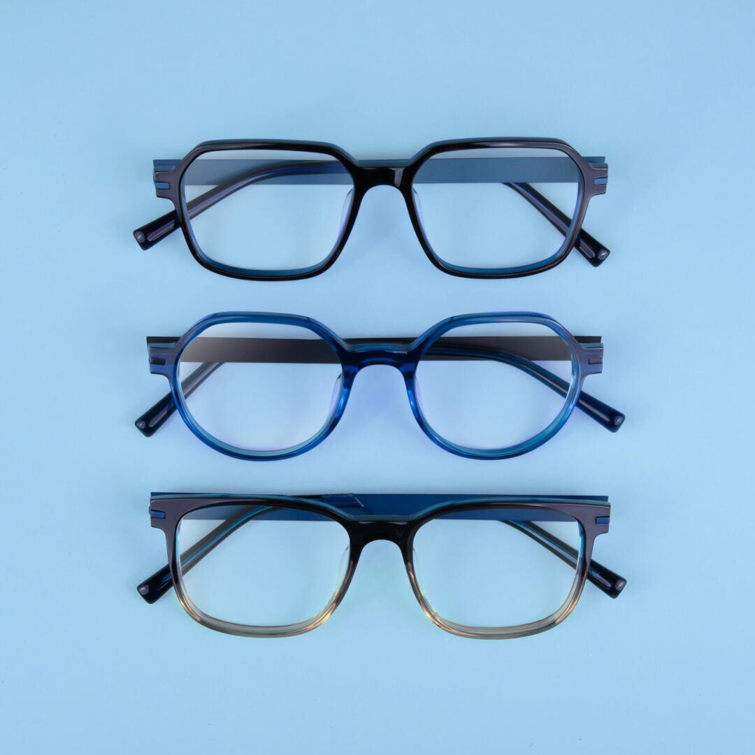 Three pairs of eyeglasses with different frame shapes and colors are arranged vertically on a light blue background. The top pair is black and rectangular, the middle pair is blue and round, and the bottom pair is black and brown gradient.