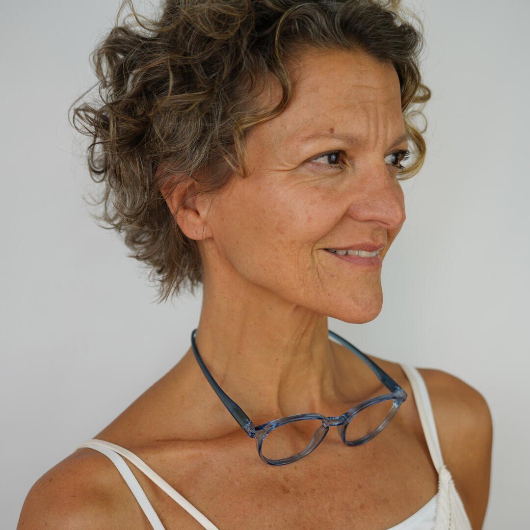 A woman with short, curly brown hair, wearing a white tank top and glasses around her neck, looks to the side and smiles against a plain white background.