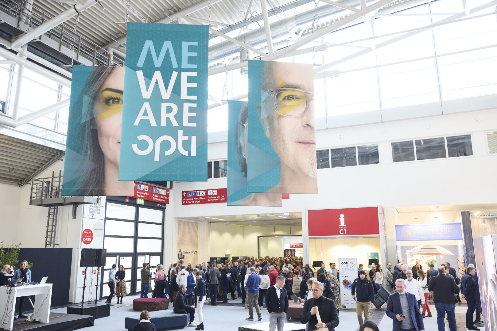 Große Banner mit Gesichtern und der Aufschrift „ME WE ARE opti“ hängen über einer belebten, modernen Messehalle, die mit Menschen auf einer Veranstaltung gefüllt ist. Im Hintergrund sind Messestände und Informationsstände zu sehen.