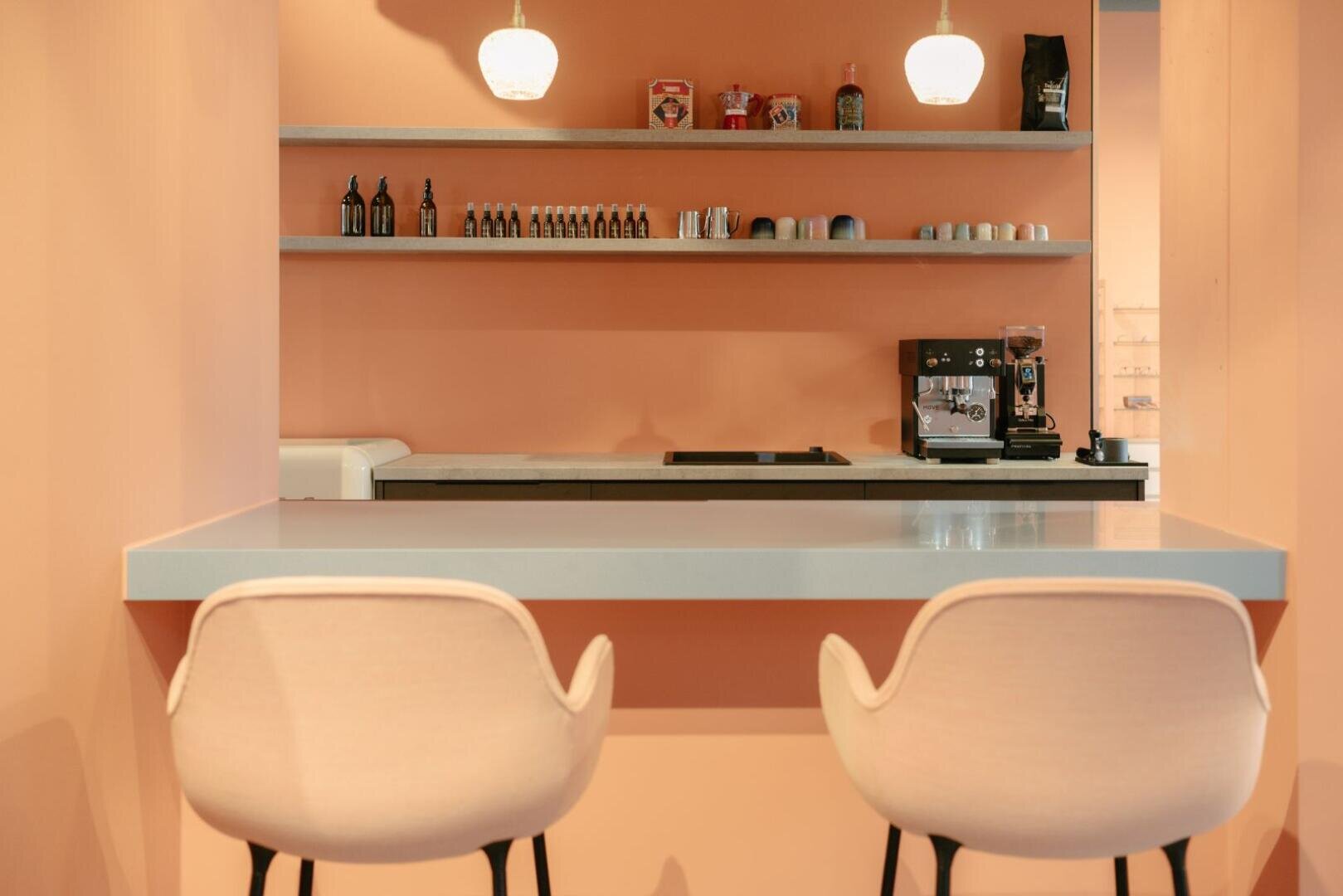 Ein modernes Café mit pfirsichfarbenen Wänden, zwei weißen Stühlen an einem Tresen, darüber Hängeleuchten und Regalen mit Flaschen, Tassen und einer Kaffeemaschine im Hintergrund.