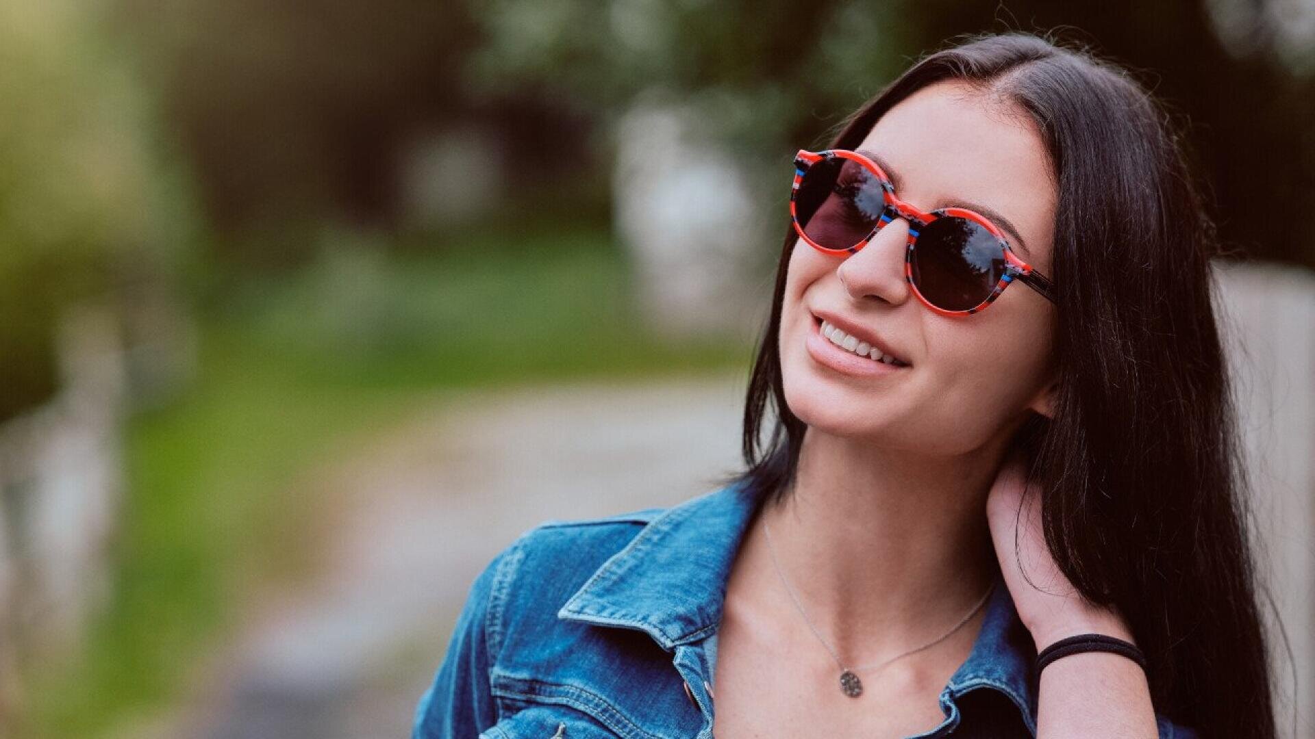A young woman with long dark hair, wearing red sunglasses and a denim jacket, smiles while touching her hair outdoors on a blurred, green background.