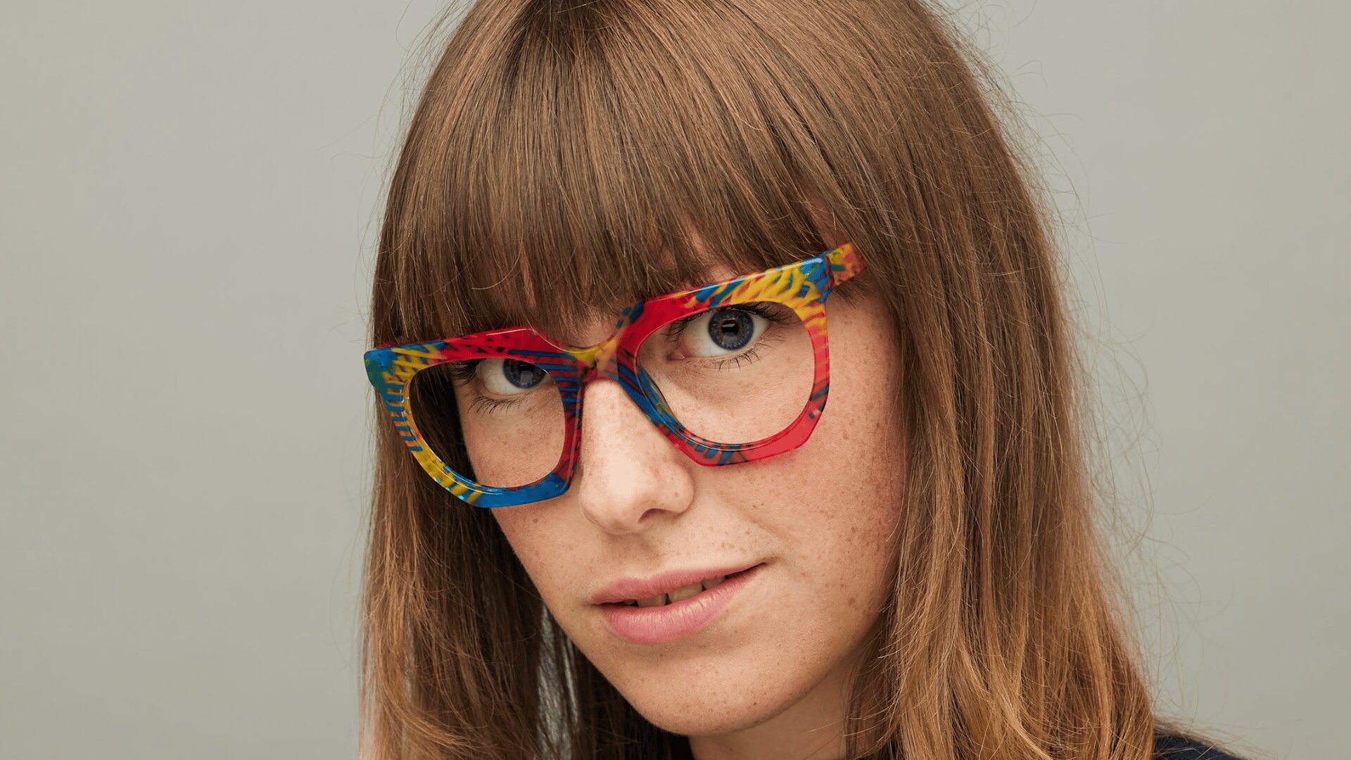 Eine Frau mit langen braunen Haaren und Pony trägt eine bunte, gemusterte Brille und blickt in die Kamera, die vor einem schlichten grauen Hintergrund steht.