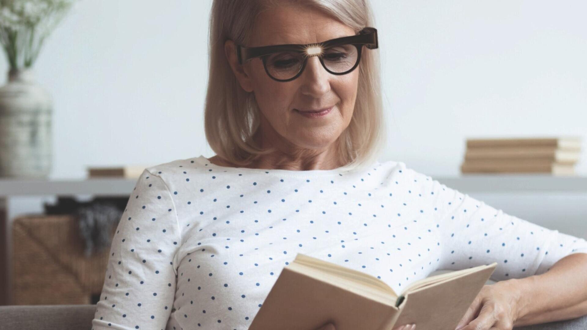 Older woman with shoulder-length blonde hair and black-rimmed glasses sits on a couch, smiling slightly while reading a book. She wears a white shirt with blue polka dots. Blurred shelves and decor are in the background.