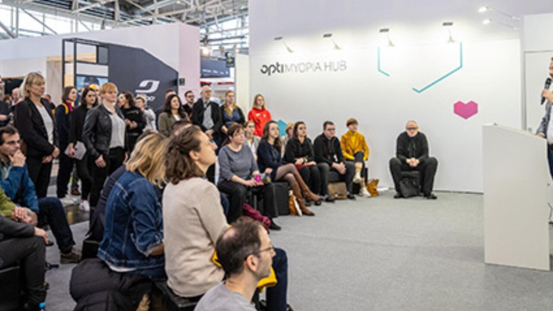 A group of people sit and stand attentively facing a speaker at an indoor event or conference, with a white display wall and a “MOPA HUB” sign in the background.