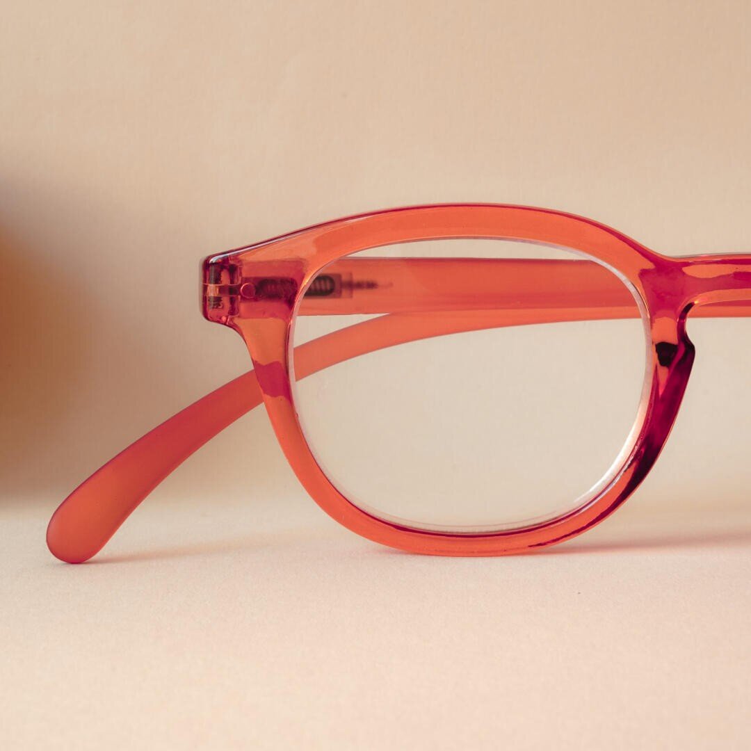 A close-up of a red spectacle frame with clear lenses resting on a light beige surface and a light beige background.