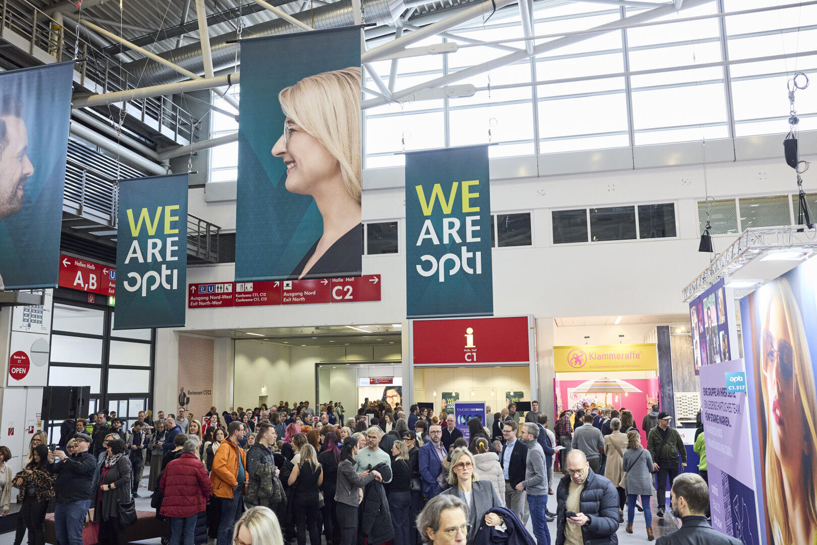 Eine große Menschenmenge versammelt sich in einem hell erleuchteten Kongresszentrum mit Transparenten mit der Aufschrift „WE ARE opti“ und lächelnden Gesichtern. Der Raum hat hohe Decken, große Fenster und über der Menge hängen Wegweiser.
