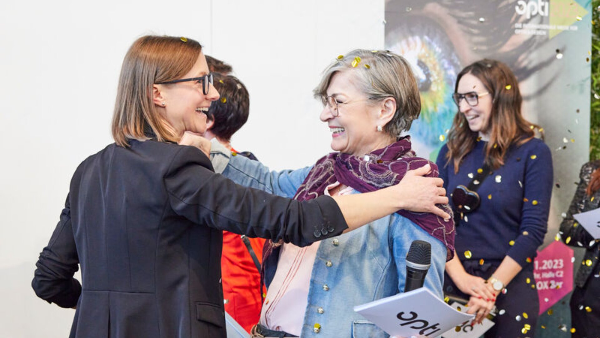 Zwei Frauen lächeln und umarmen sich bei einer Veranstaltung. Andere stehen lächelnd im Hintergrund. Eine Frau hält ein Mikrofon und ein Programm. Um sie herum regnet Konfetti. Hinter ihnen ist ein buntes Plakat teilweise sichtbar.