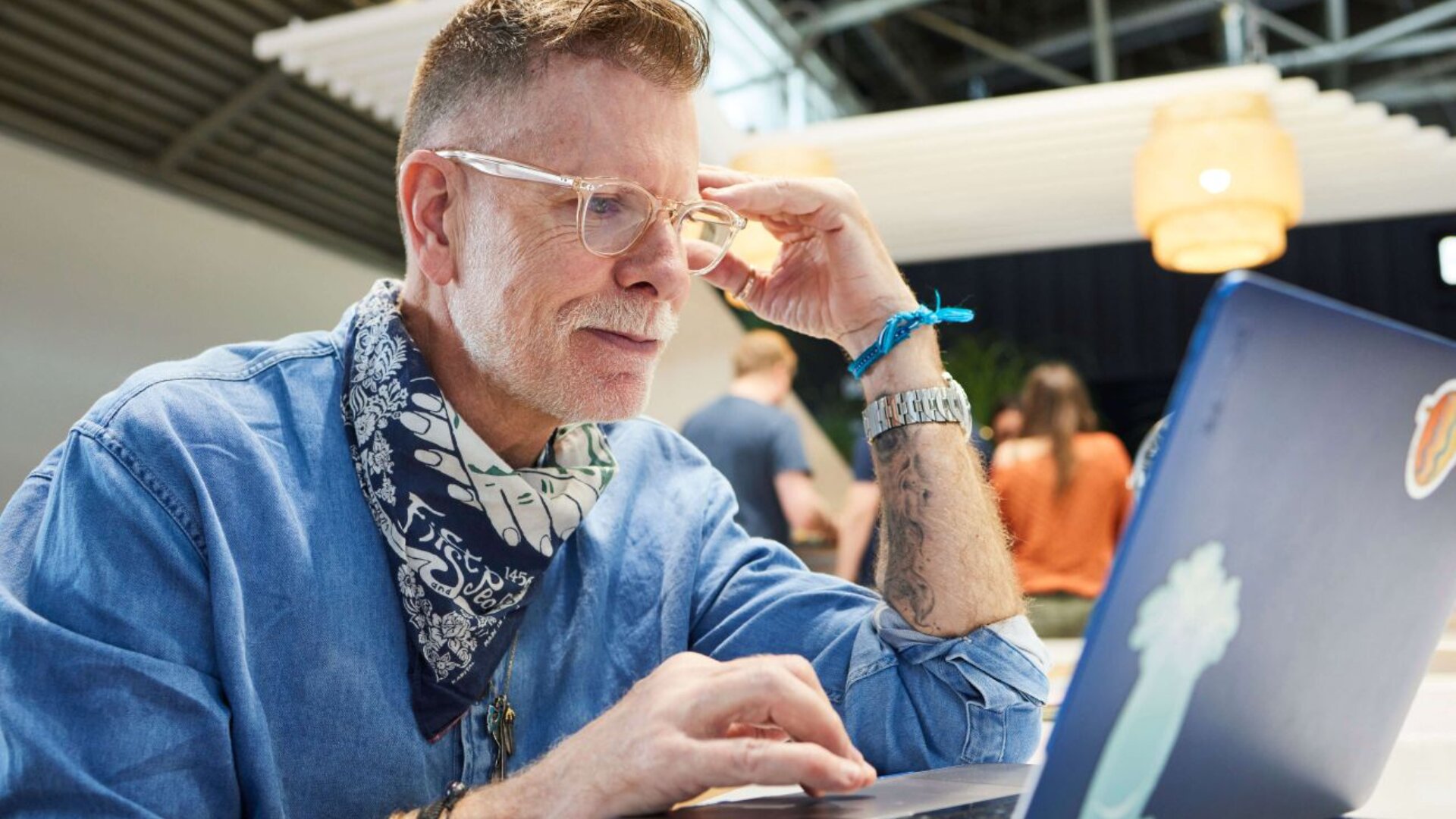 Ein älterer Mann mit Brille und Bandana sitzt an einem Tisch und konzentriert sich auf seinen Laptop. Er trägt ein Jeanshemd und hat Tattoos am Arm. Im Hintergrund sind Menschen und moderne Beleuchtung verschwommen.