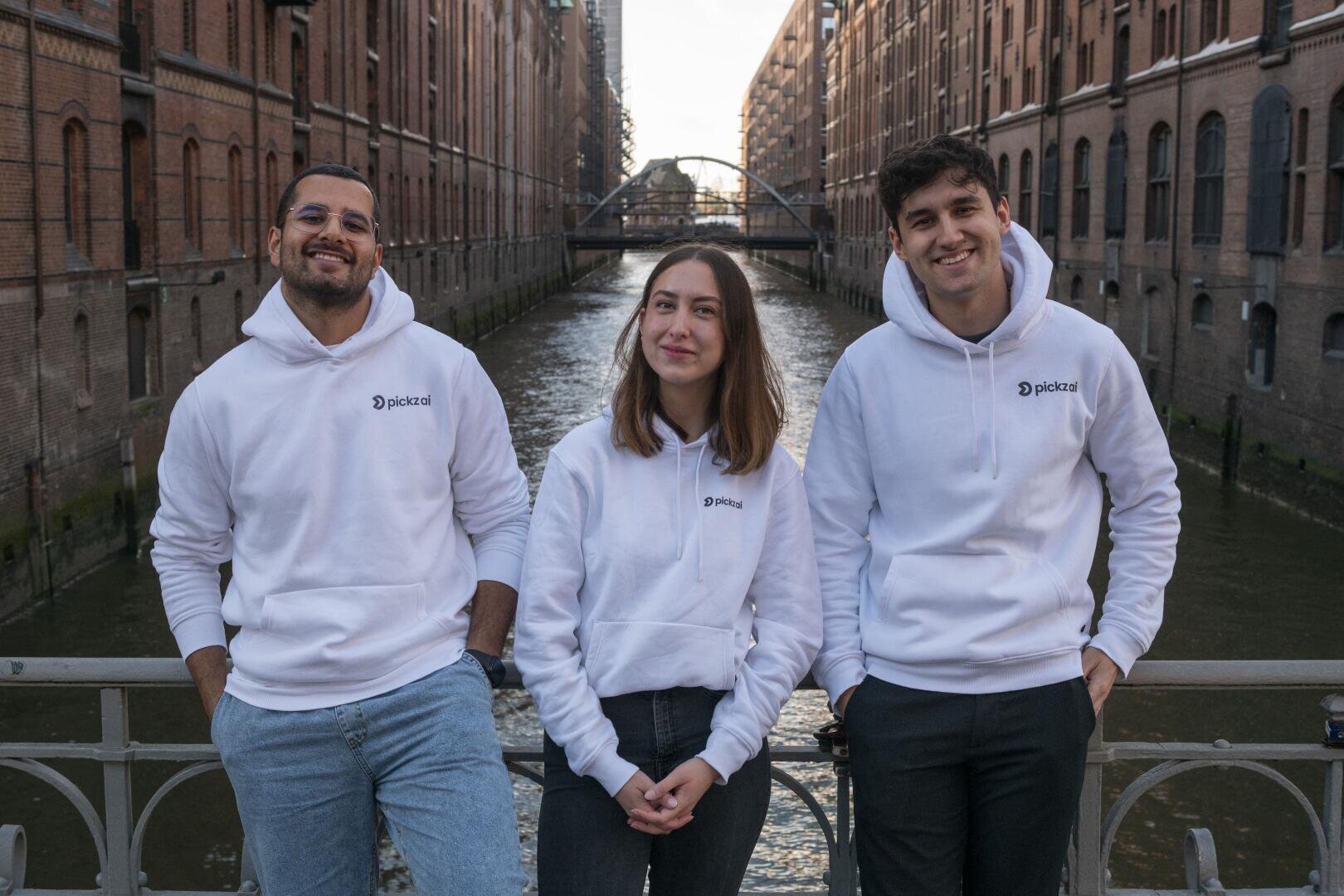 Die drei Gründer:in von pickz ai, die weiße Kapuzenpullis mit Logo tragen, stehen auf einer Brücke in Hamburg über einen Kanal mit Backsteinbauten auf beiden Seiten. Sie lächeln und posieren für das Foto.