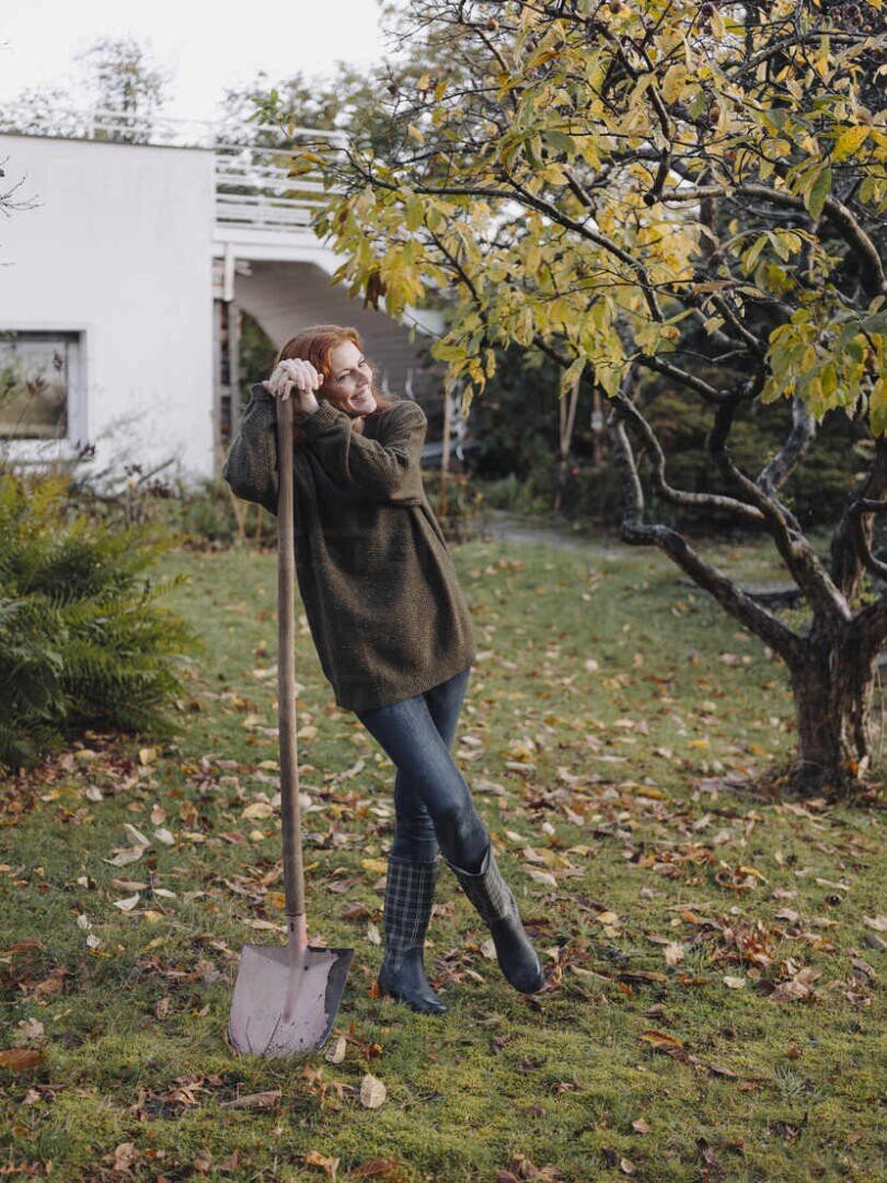 Eine Frau in Pullover und Stiefeln steht lächelnd im Gras und stützt sich auf eine Schaufel in einem Garten mit Herbstlaub, in der Nähe eines Baums und eines weißen Hauses.