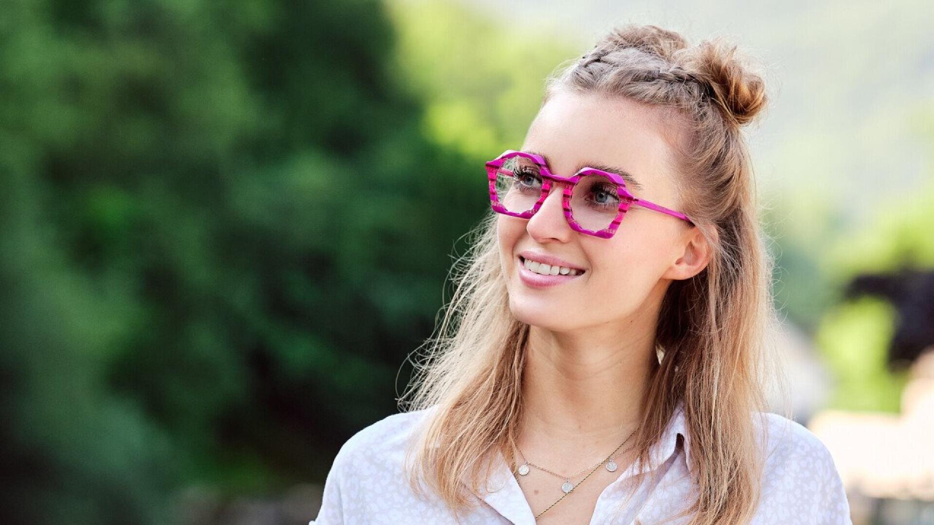 A smiling woman with light brown hair styled in two buns wears bright pink, geometric glasses and a white shirt, standing outdoors with a blurred green background.
