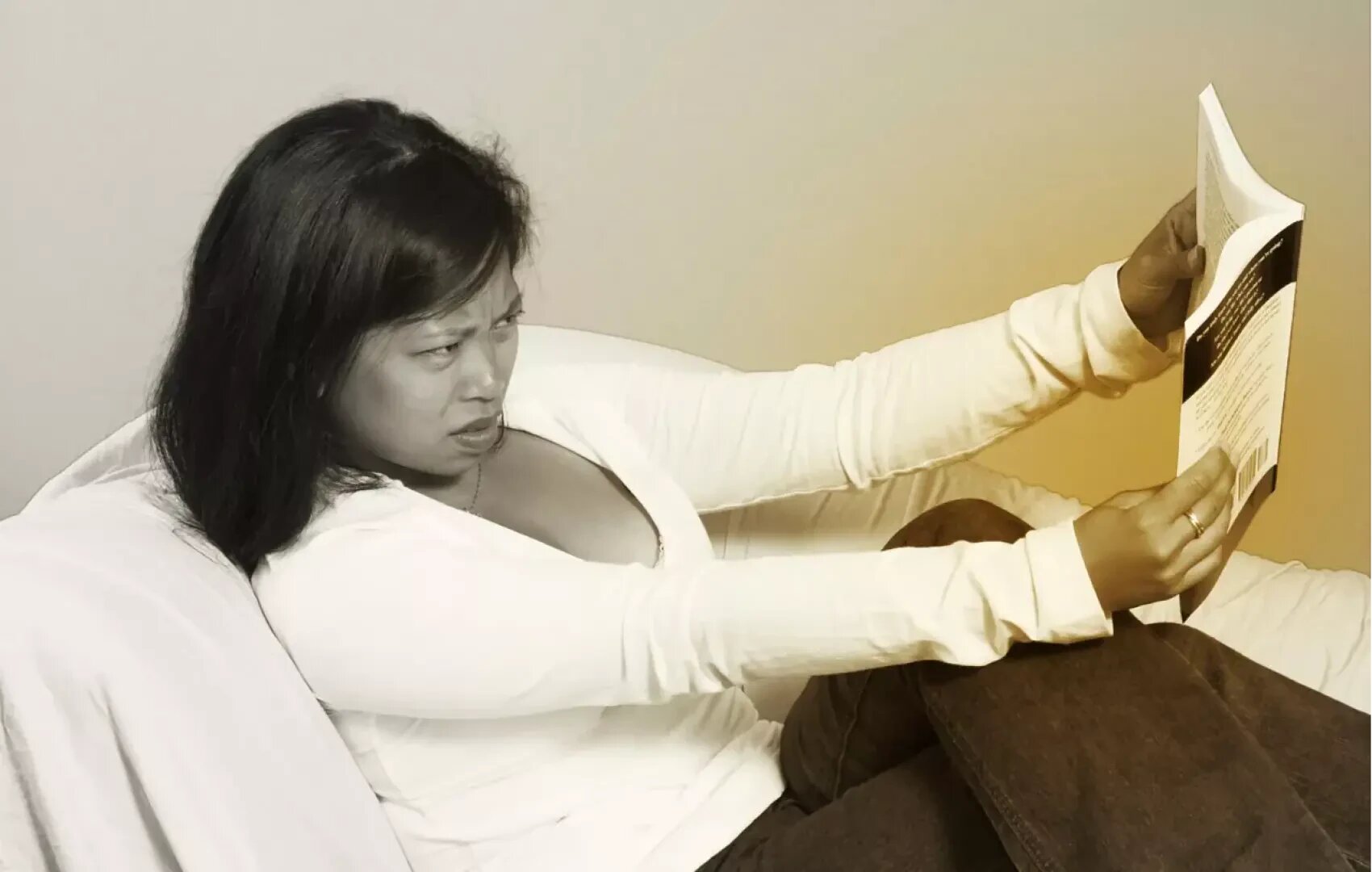 Eine Frau sitzt auf einem Sofa, hält ein Buch auf Armeslänge von sich und blinzelt verwirrt oder frustriert darauf. Sie trägt ein legeres weißes Top und Jeans. Der Hintergrund ist neutral.