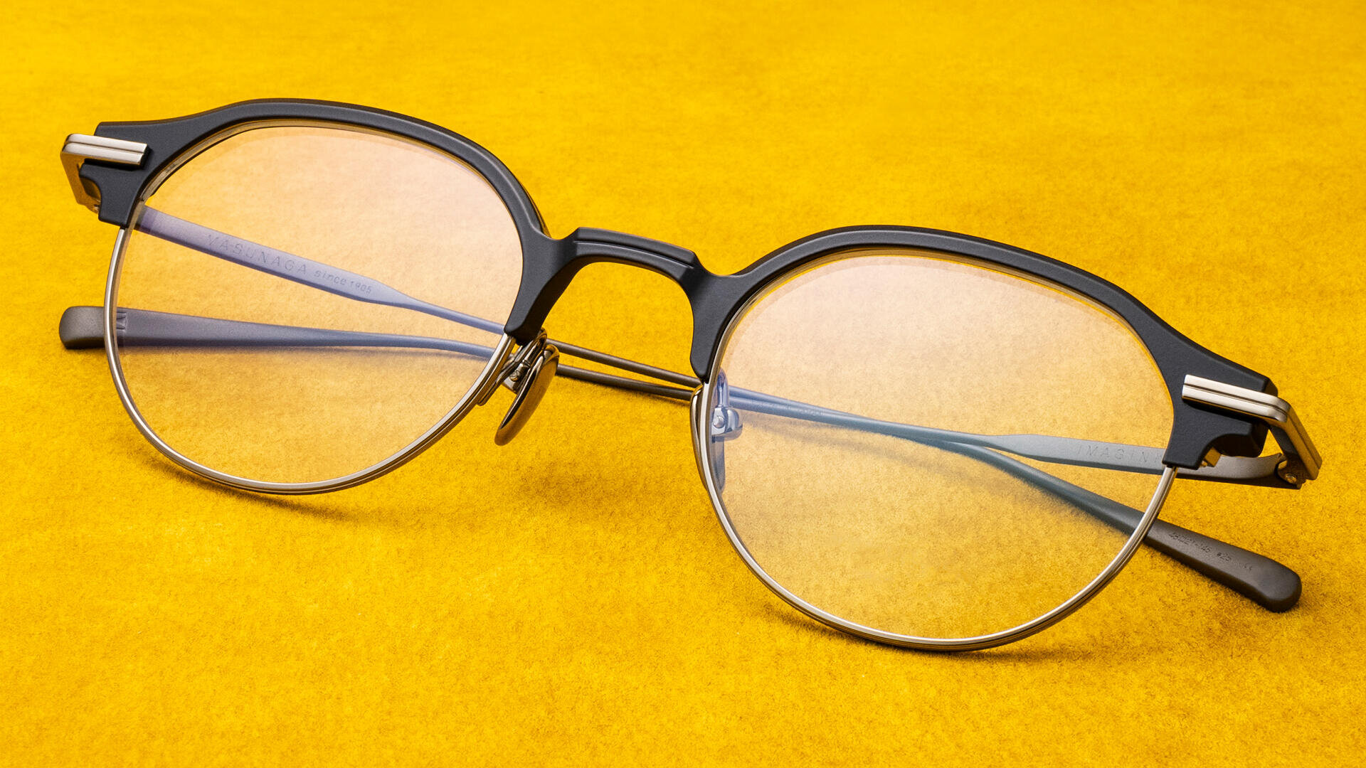 Auf einem leuchtend gelben Hintergrund ist eine runde, schwarz-silberne Brille mit klaren Gläsern abgebildet.