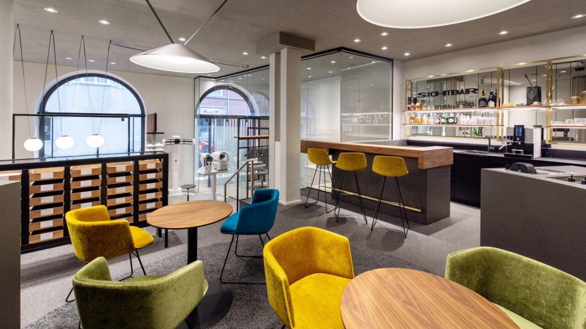 Modern café with round wooden tables, colorful upholstered chairs in yellow, green, and blue, a sleek bar area with wooden counter and barstools, large arched windows, and contemporary lighting.