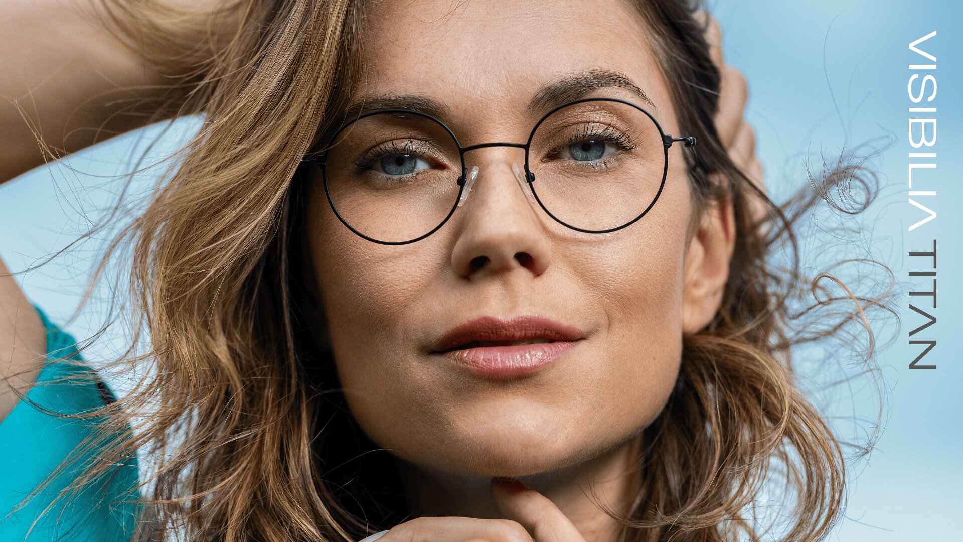 Eine Frau mit gewelltem braunem Haar und runder Brille blickt selbstbewusst in die Kamera vor einem blauen Hintergrund. Der Text VISIBILIA TITAN erscheint vertikal auf der rechten Seite.