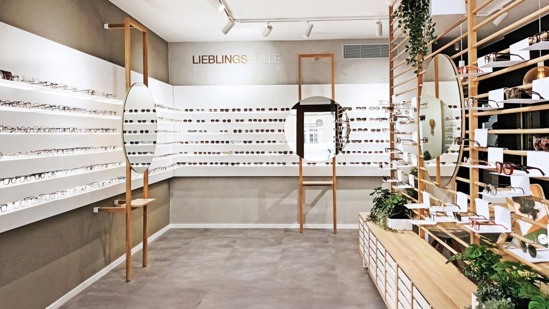 A modern eyewear store with white shelves lined with various eyeglasses, wooden accents, two round mirrors, and green plants decorating the interior. The word LIEBLINGS is displayed on the back wall.