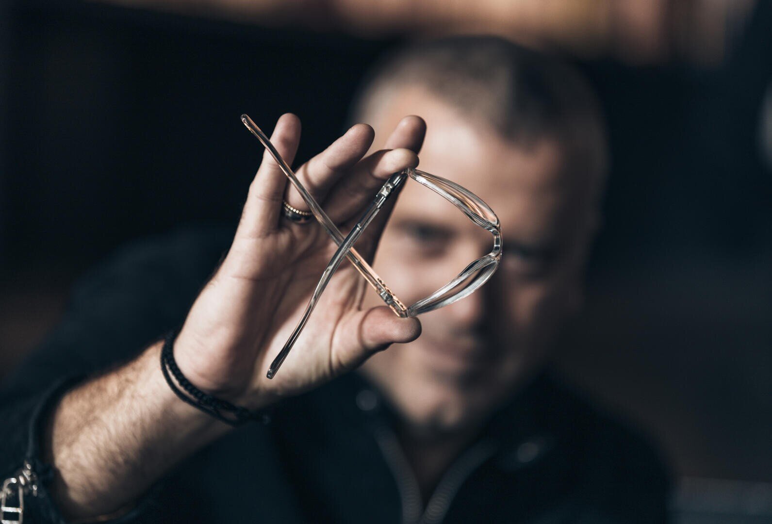 Eine Person hält eine Brille an den Schläfen hoch. Die Brille ist scharf abgebildet, ihr Gesicht ist im Hintergrund verschwommen. Die Person trägt ein dunkles Hemd und ein Armband.