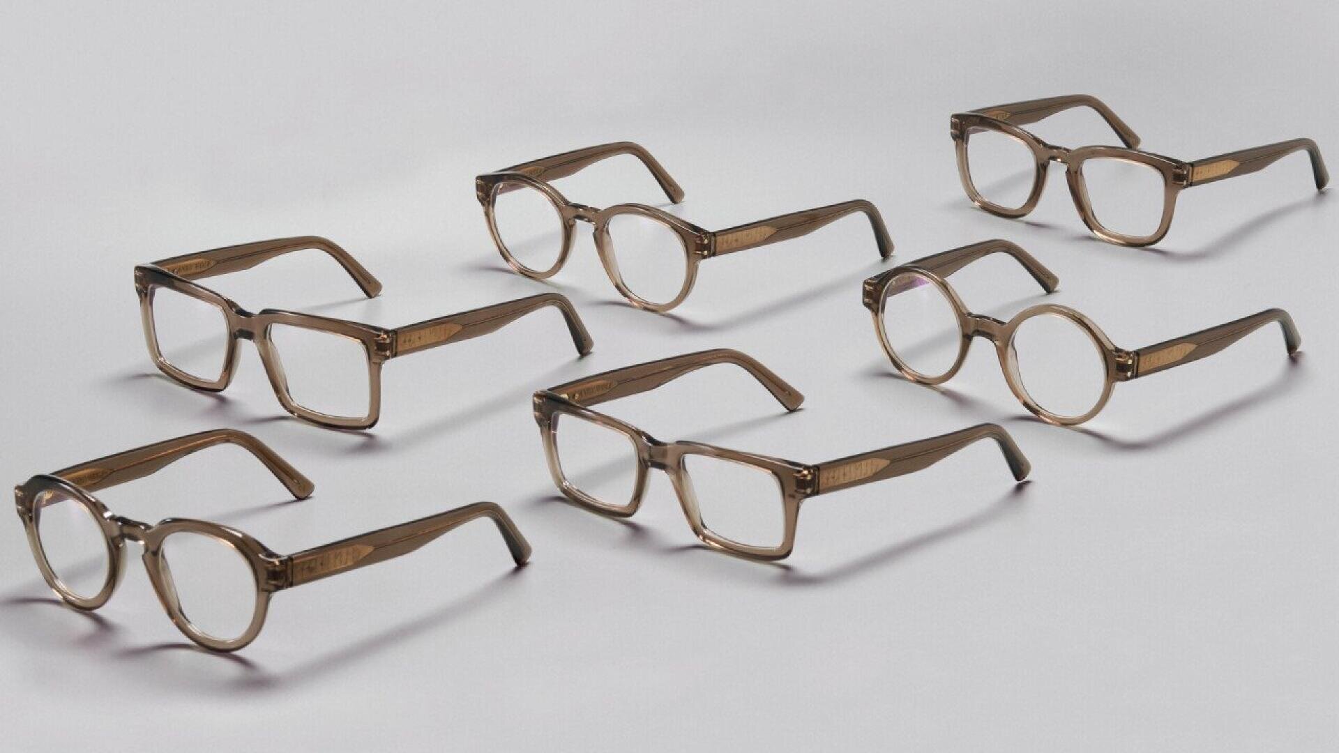 Seven pairs of light brown eyeglasses with clear lenses are arranged on a white surface. Most of the frames are rectangular, while two pairs have round frames.