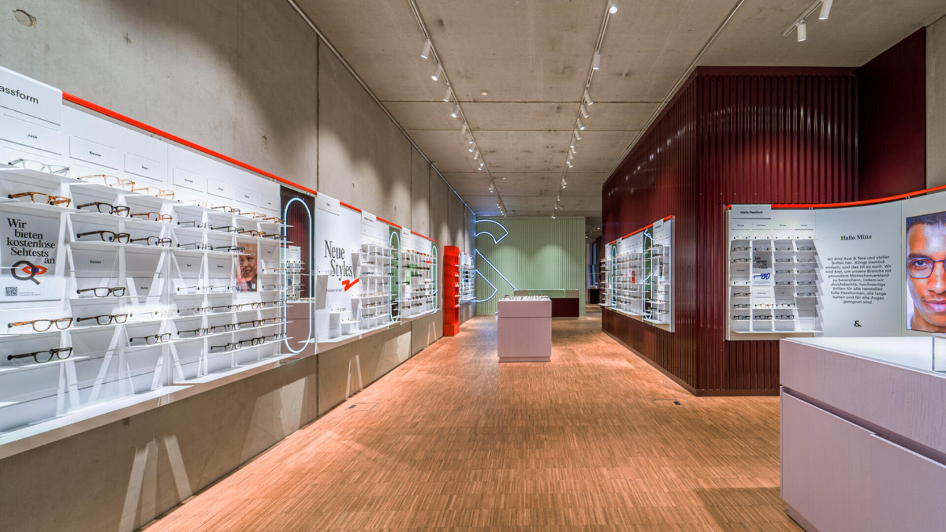 Ein modernes Brillengeschäft mit Brillenpräsentationen auf weißen Regalen an beiden Wänden, Holzboden und heller Deckenbeleuchtung. Ein freistehendes Display und Informationsplakate sind überall im Raum zu sehen.