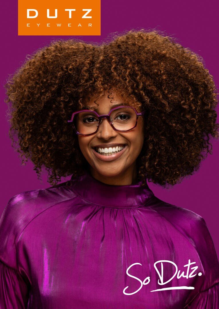 A smiling woman with curly hair wears purple glasses and a shiny purple top, standing in front of a purple background. The image has Dutz Eyewear and So Dutz. written on it.