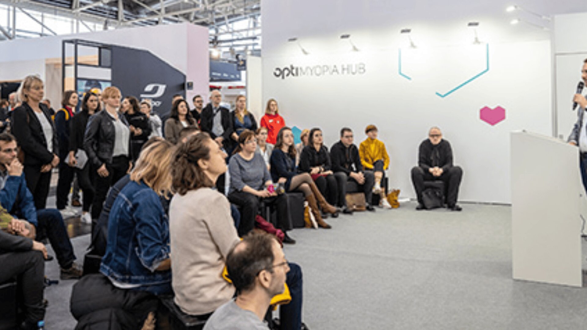 Eine Gruppe von Personen sitzt einem Redner gegenüber, der an einem Podium steht und in einem Veranstaltungsraum mit der Bezeichnung OPTIMORA HUB präsentiert. Das Publikum scheint aufmerksam zu sein, und die Umgebung ist modern und gut beleuchtet.