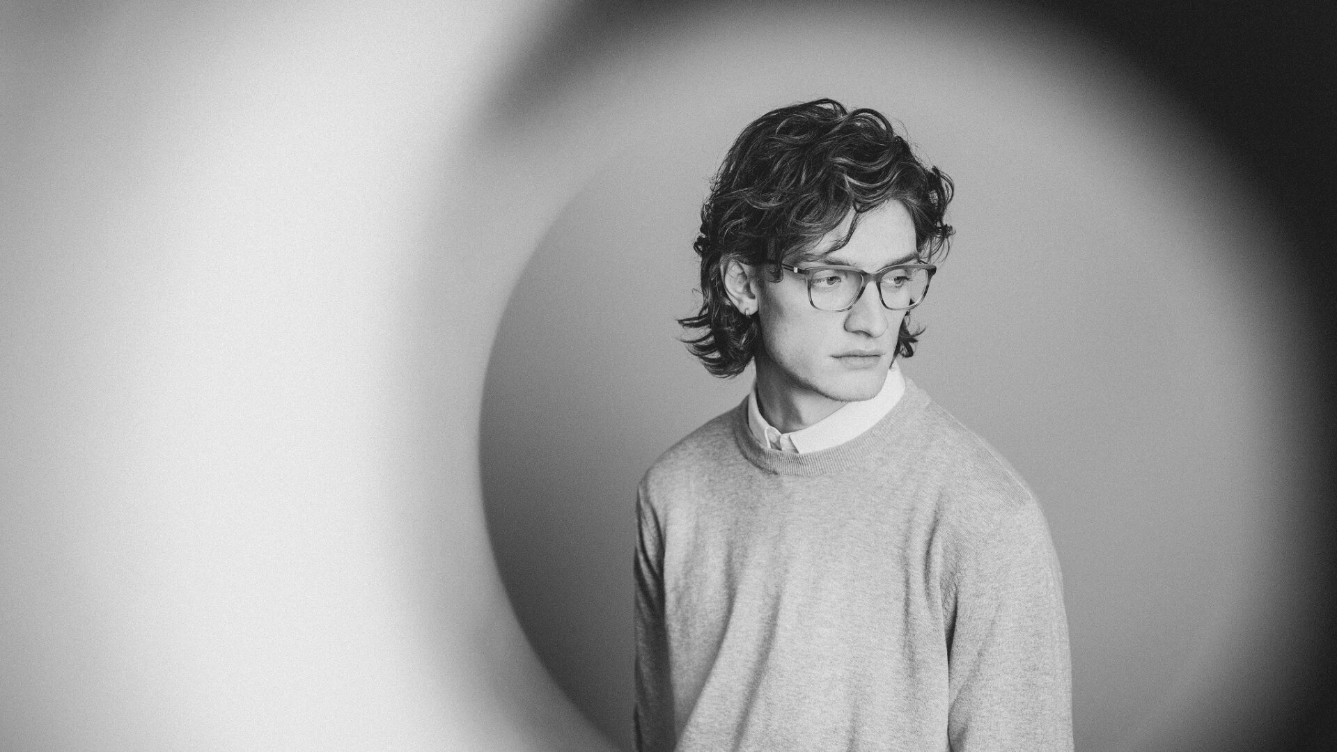 Schwarz-Weiß-Foto eines jungen Mannes mit welligem Haar und Brille. Er trägt einen leichten Pullover über einem Hemd mit Kragen und blickt nachdenklich zur Seite. Der Hintergrund zeigt weiche, kreisförmige, unscharfe Muster.