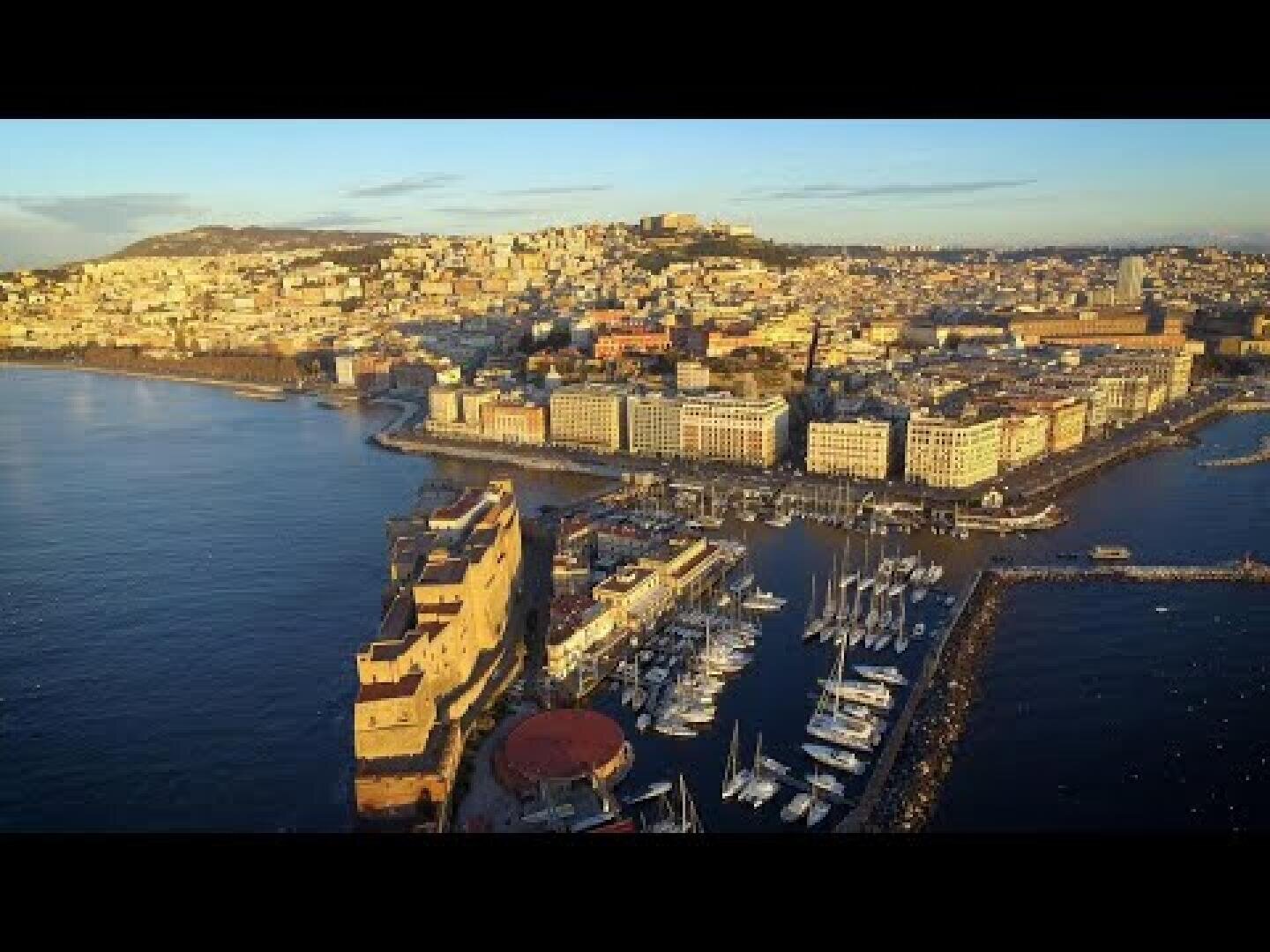 Aerial view of Naples, Italy, showing a marina with docked boats, historic buildings along the waterfront and the city stretching over the hills under a clear sky at sunset.