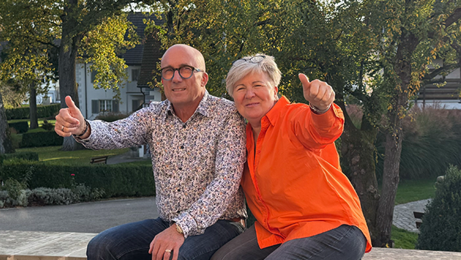 Ein Mann und eine Frau sitzen draußen auf einem Felsvorsprung. Beide lächeln und zeigen mit dem Daumen nach oben. Der Mann trägt eine Brille und ein gemustertes Hemd; die Frau trägt ein orangefarbenes Hemd. Im Hintergrund sind grüne Bäume und Gebäude zu sehen.
