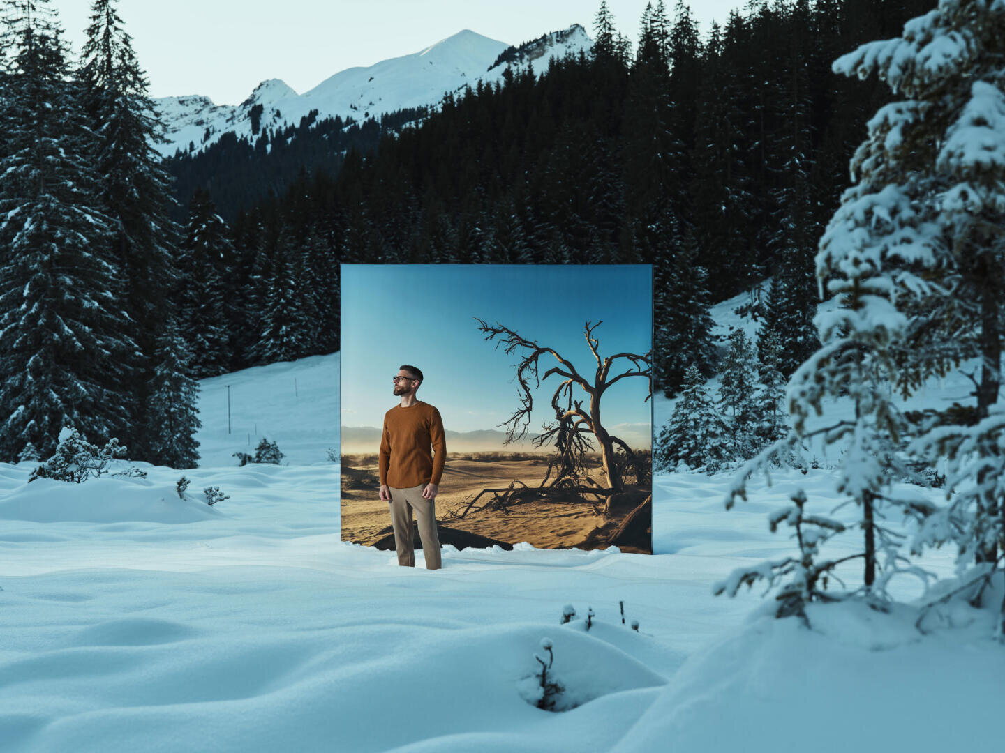 Ein Mann steht im tiefen Schnee zwischen Kiefern und Bergen. Hinter ihm ist ein großes Foto einer trockenen, kargen Landschaft mit einem abgestorbenen Baum zu sehen, das einen starken Kontrast zur verschneiten Umgebung bildet.