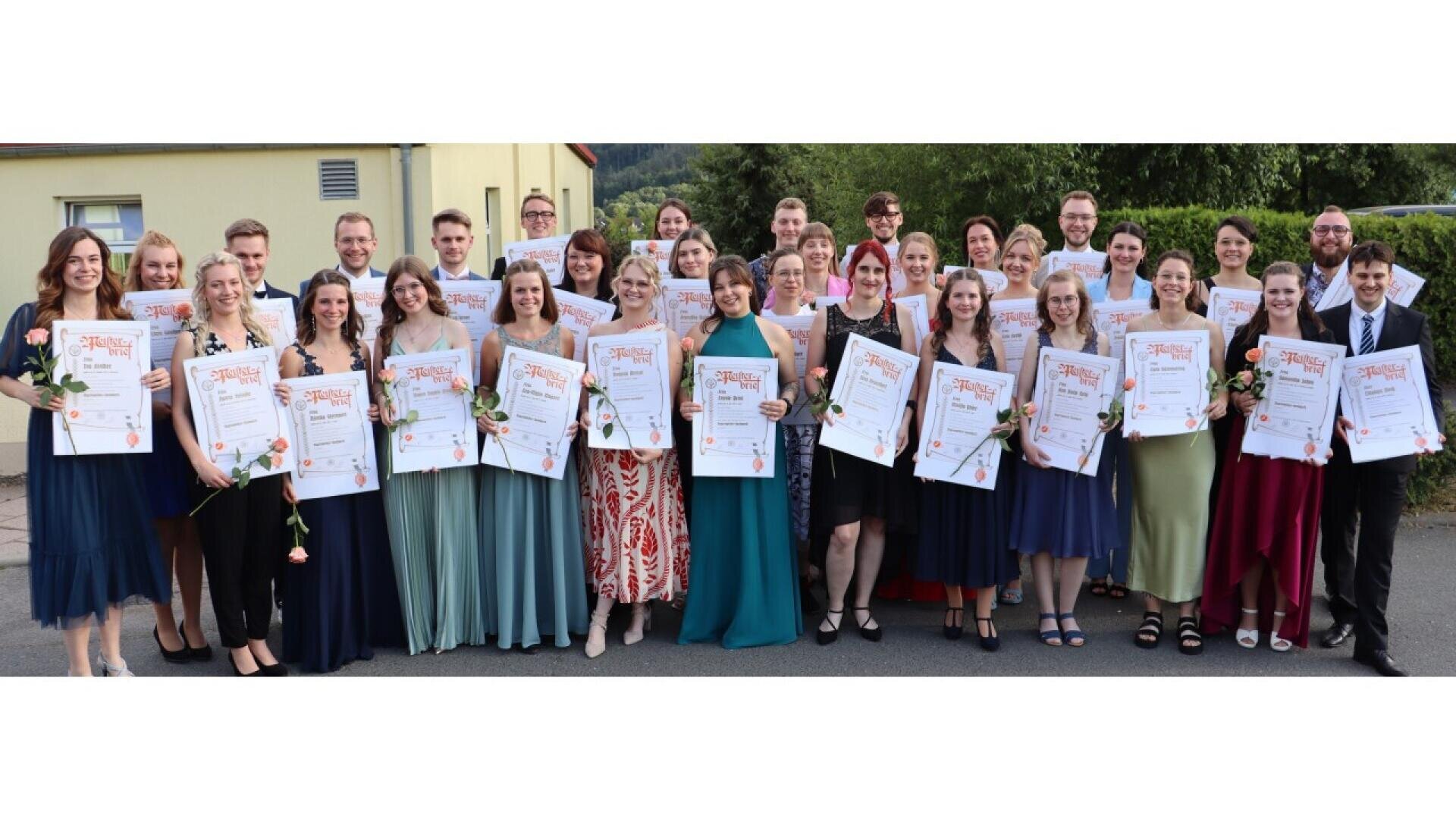A large group of young adults, dressed formally, stand outdoors holding certificates and single roses, smiling at the camera. Trees and a light-yellow building are visible in the background.