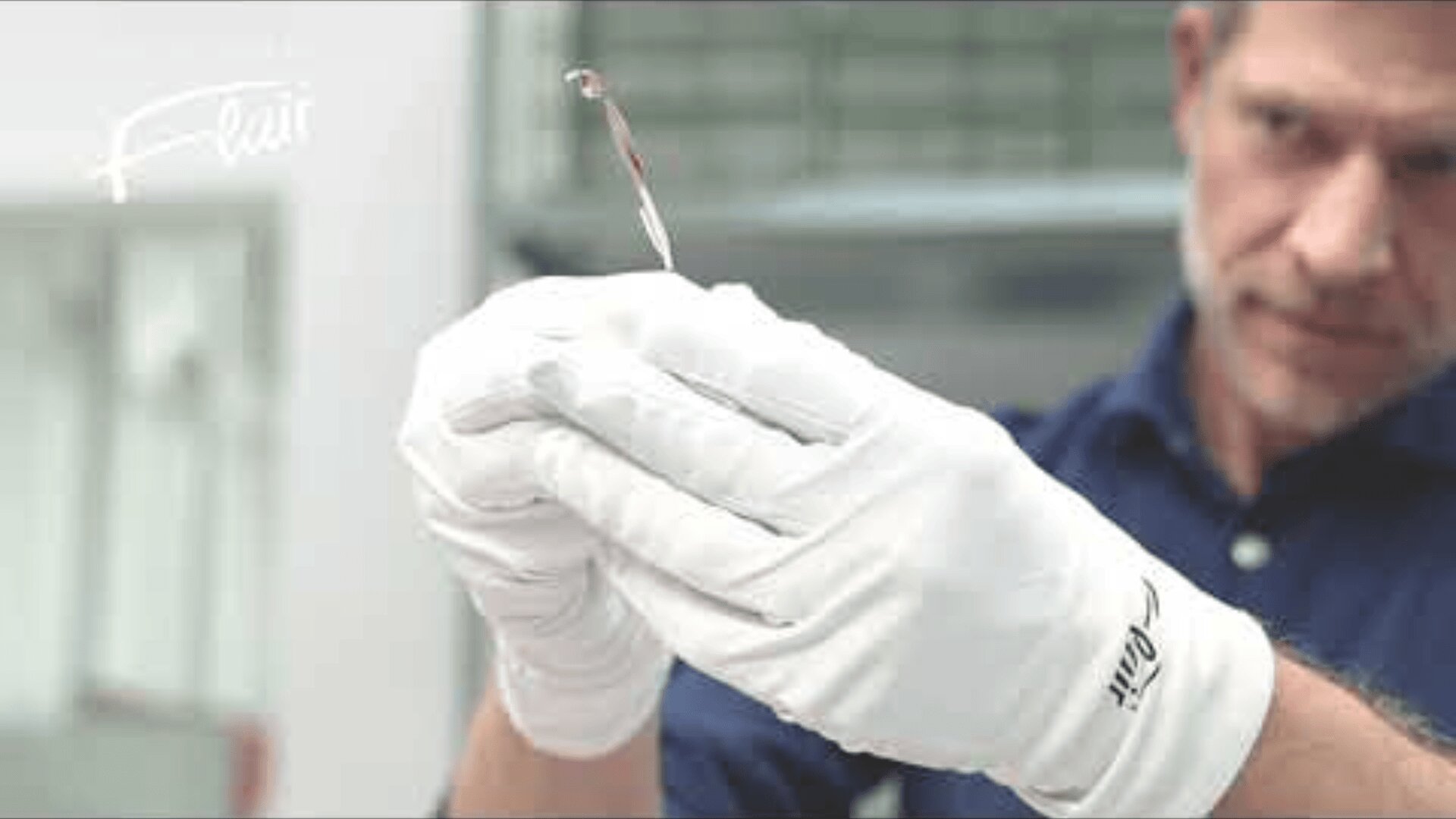 A man wearing white gloves carefully holds a thin, transparent object with a small red tip, examining it closely in a laboratory or workshop setting.