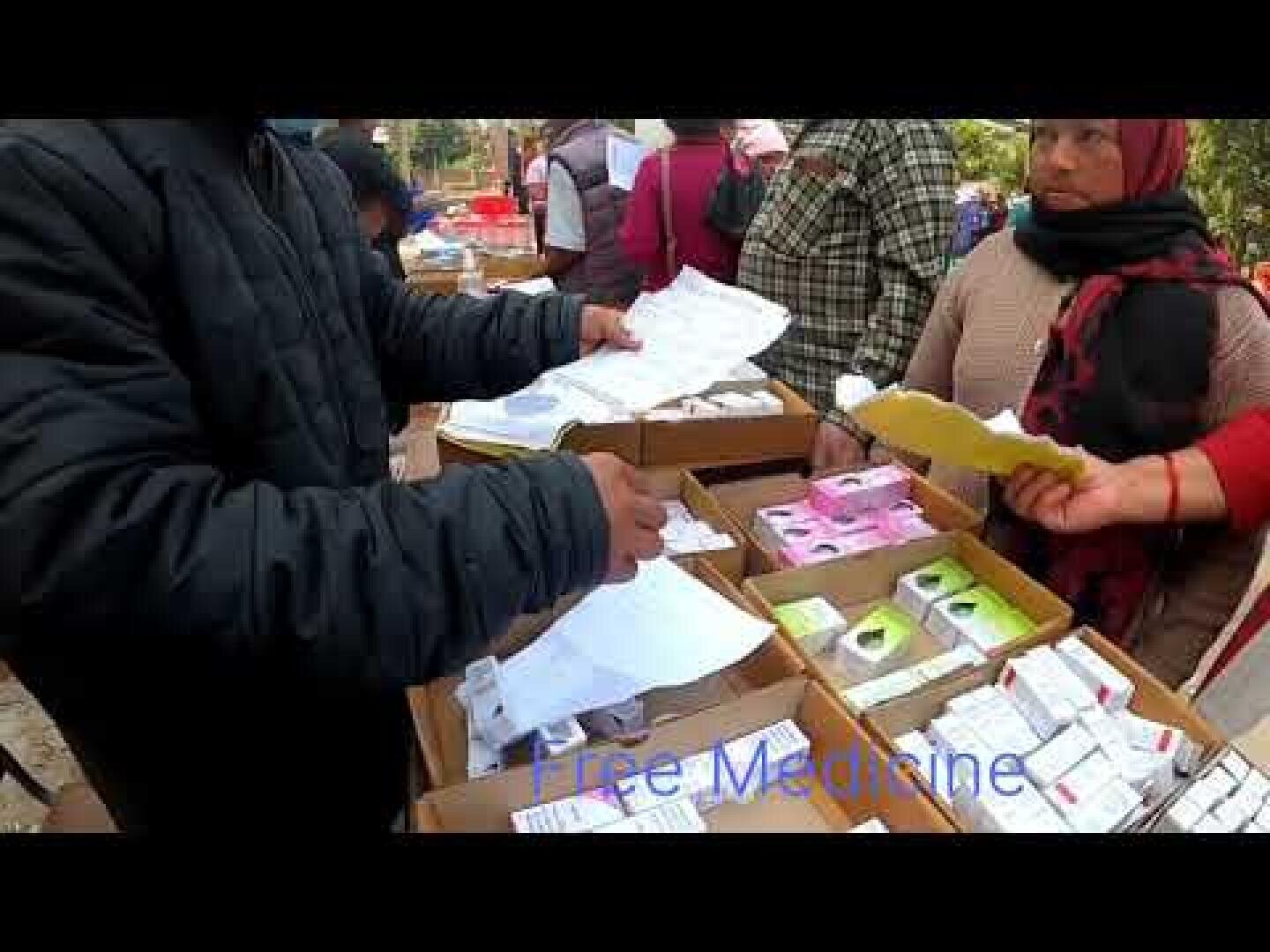 A person hands out papers near boxes of various medicines at an outdoor stall, while others stand nearby. The words Free Medicine are faintly visible at the bottom of the image.