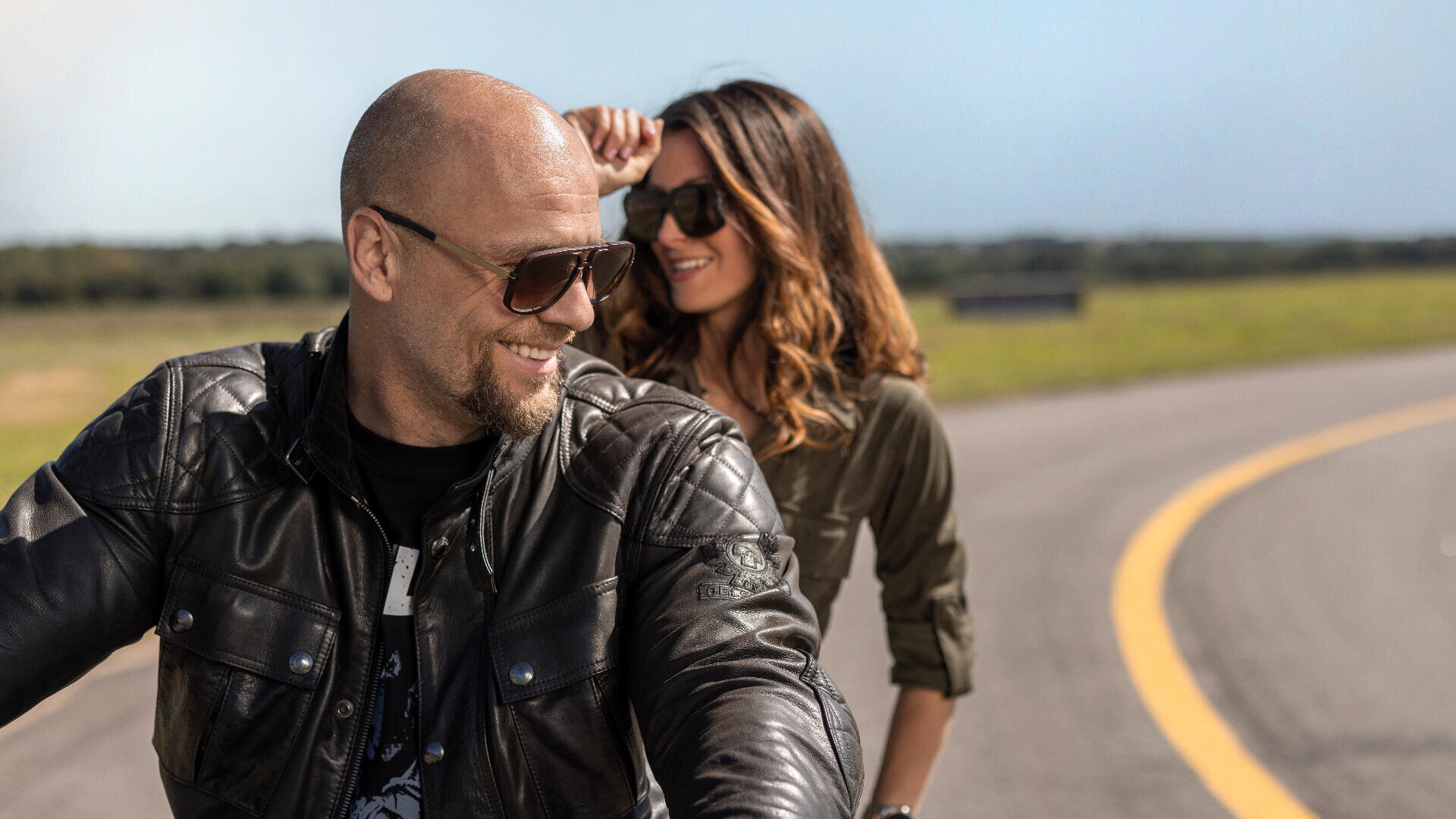 Ein Mann und eine Frau mit Sonnenbrillen und Lederjacken lächeln, während sie auf einem Motorrad auf einer leeren Straße mit klarem Himmel und offenen Feldern im Hintergrund fahren.
