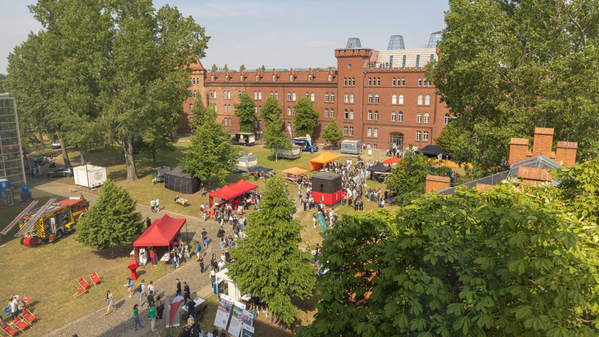 Luftaufnahme eines Open-Air-Festivals mit bunten Zelten, Essensständen und Menschen, die sich auf einer Rasenfläche vor einem großen, von Bäumen umgebenen Gebäude aus rotem Backstein versammelt haben.