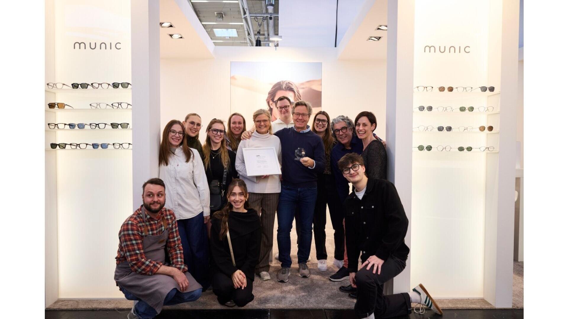A group of people pose and smile together inside a modern eyewear store, standing between displays of glasses with “MUNIC” written on the walls. One person holds a framed certificate, and another holds a glass award.