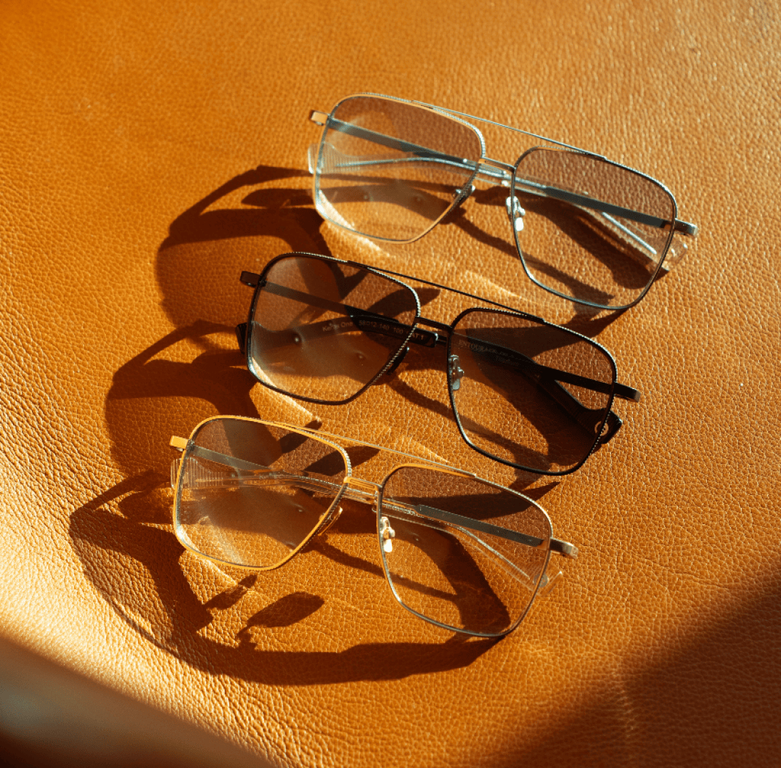Three pairs of aviator-style eyeglasses with gold, black, and silver frames are arranged in a row on a textured brown leather surface, casting distinct shadows in the sunlight.