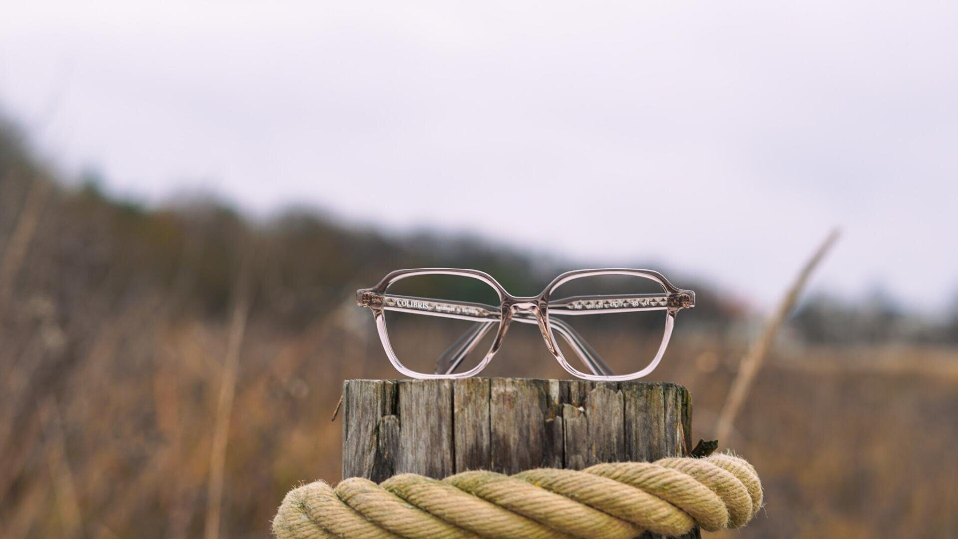 A pair of glasses rests on a weathered wooden post with a thick rope wrapped around it; the background is blurred and shows a natural environment.