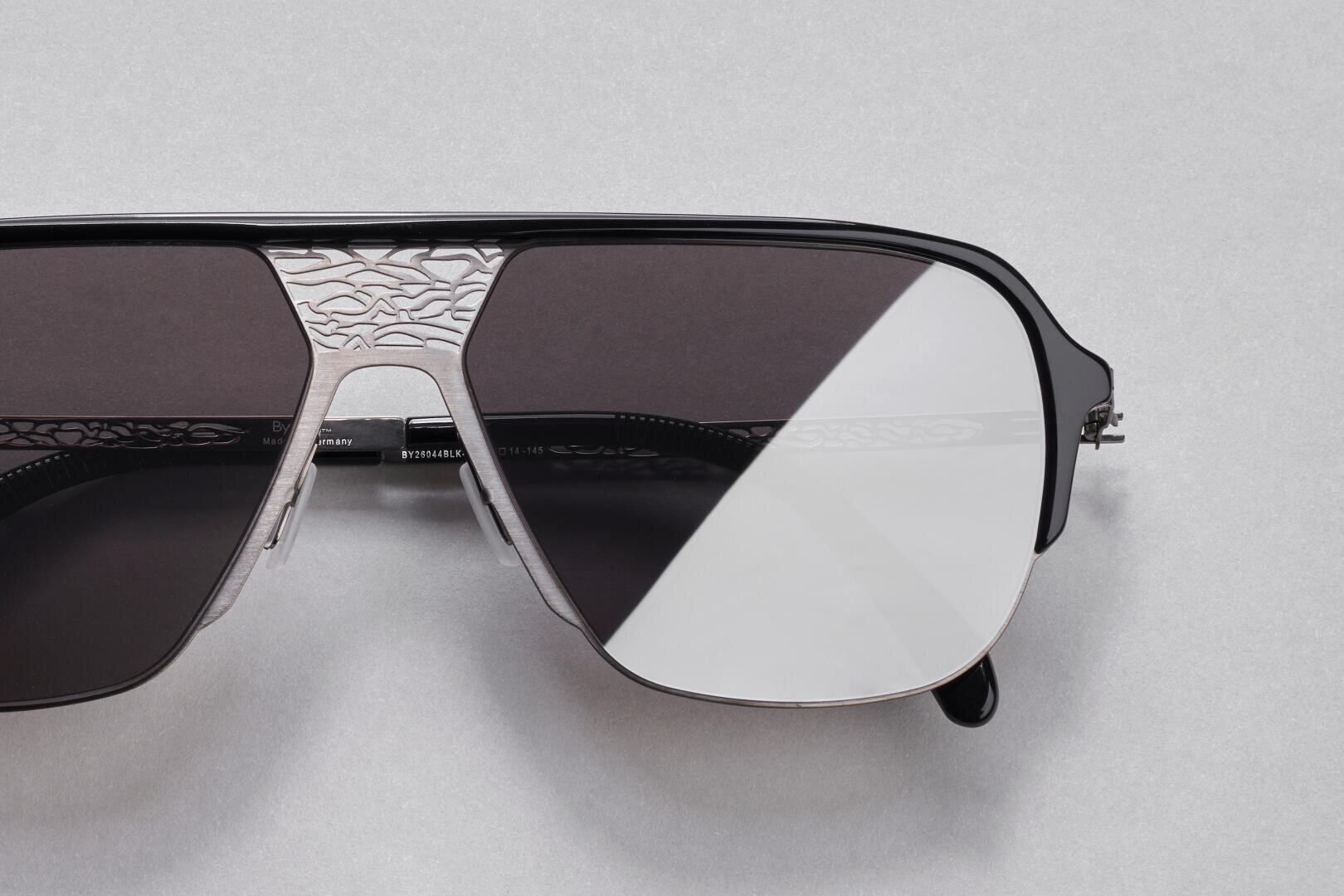 Close-up of stylish sunglasses with a silver metal frame and dark lenses, featuring an intricate engraved pattern on the nose bridge and upper rim against a light gray background.